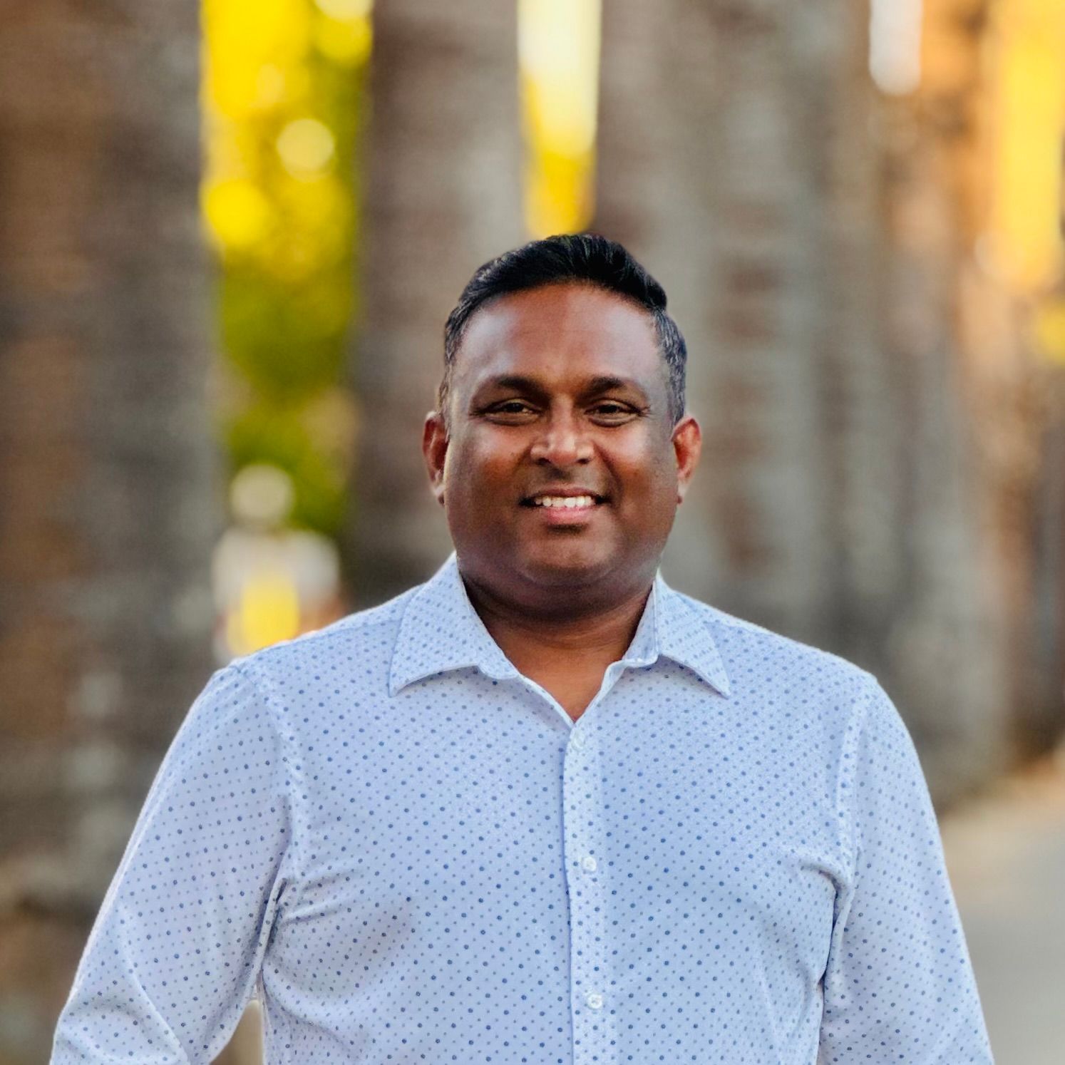 A smiling man in a white button-up shirt poses outdoors with a warm bokeh background.