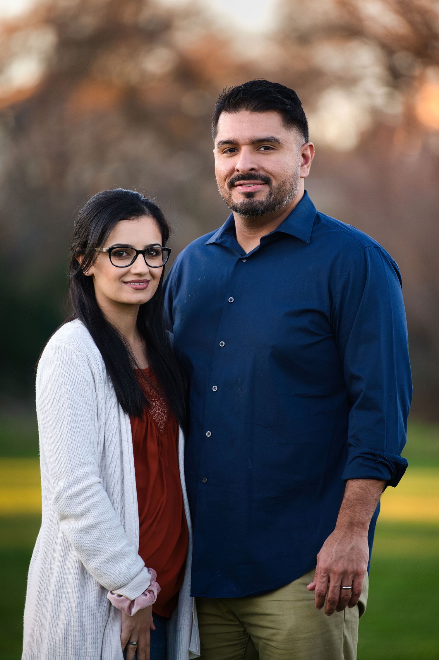 Couple posing together outdoors, woman in white cardigan, man in navy blue shirt.