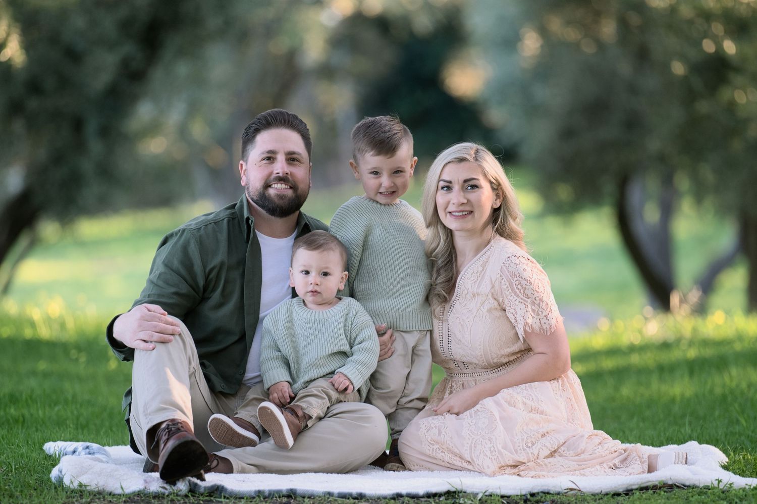 Smiling family of four sitting on blanket in park, two young boys in sage green and neutral coordinating outfits.