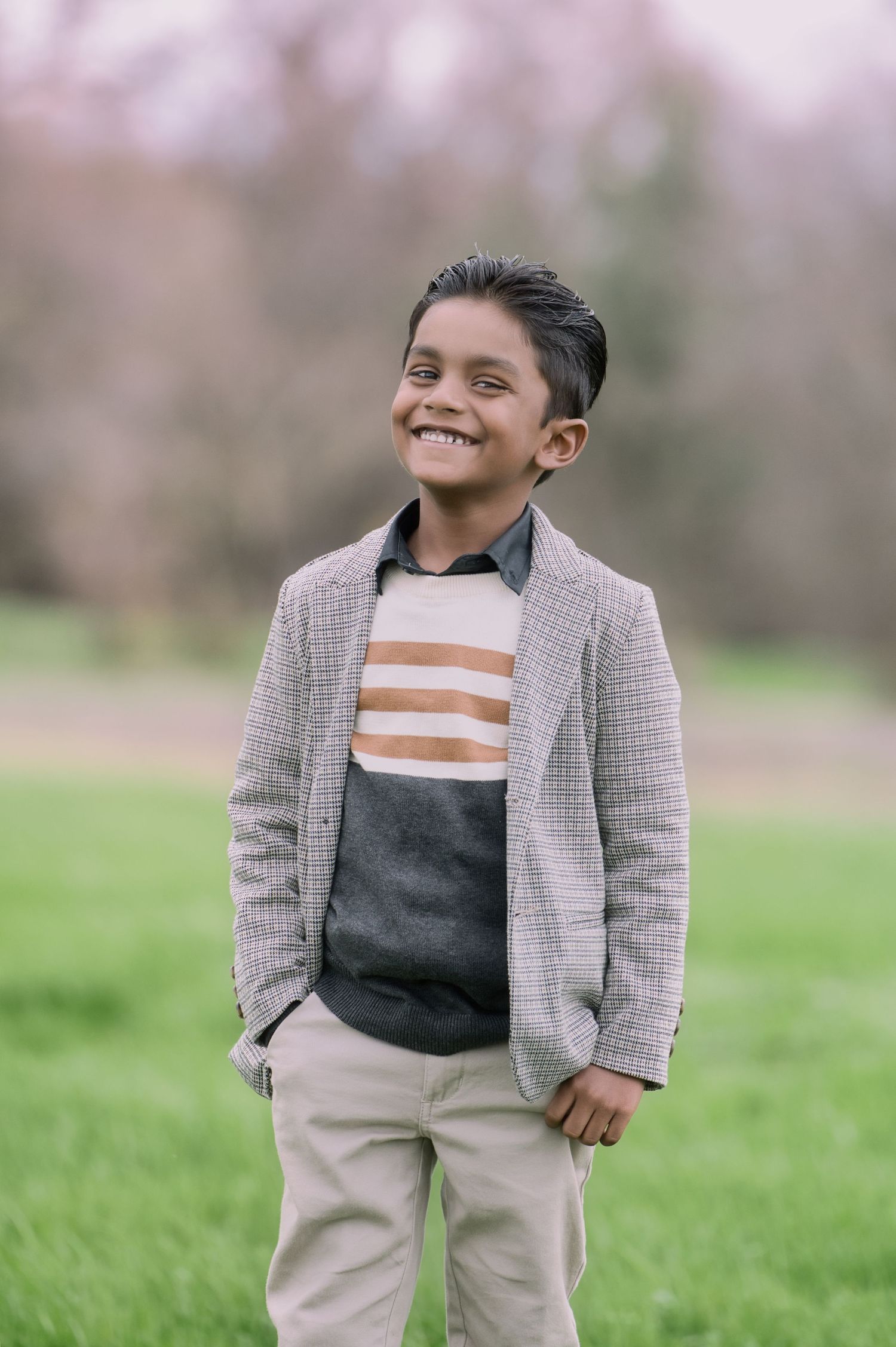 A smiling boy in a blazer and striped sweater stands on a green lawn with blossoming trees behind.