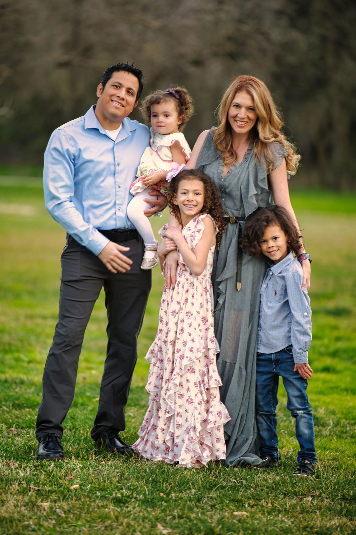 Happy family of five posing on green grass, parents with three young children in coordinated spring outfits.