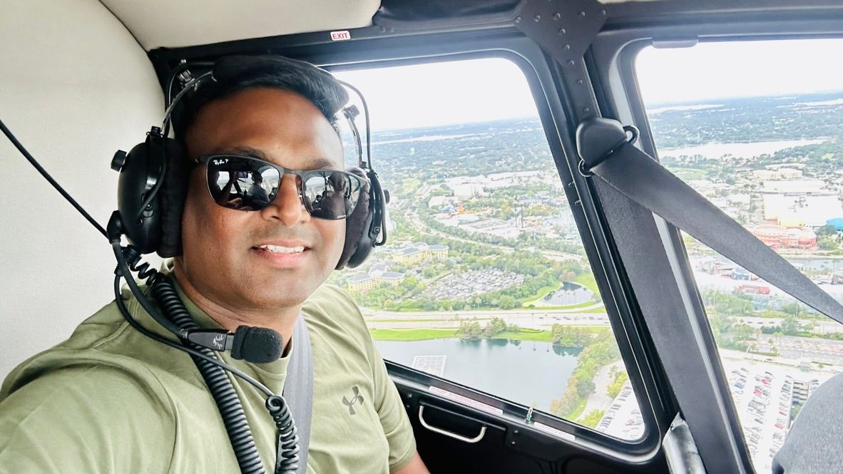 A smiling pilot in a helicopter cockpit wearing a headset and sunglasses flies over a city landscape.
