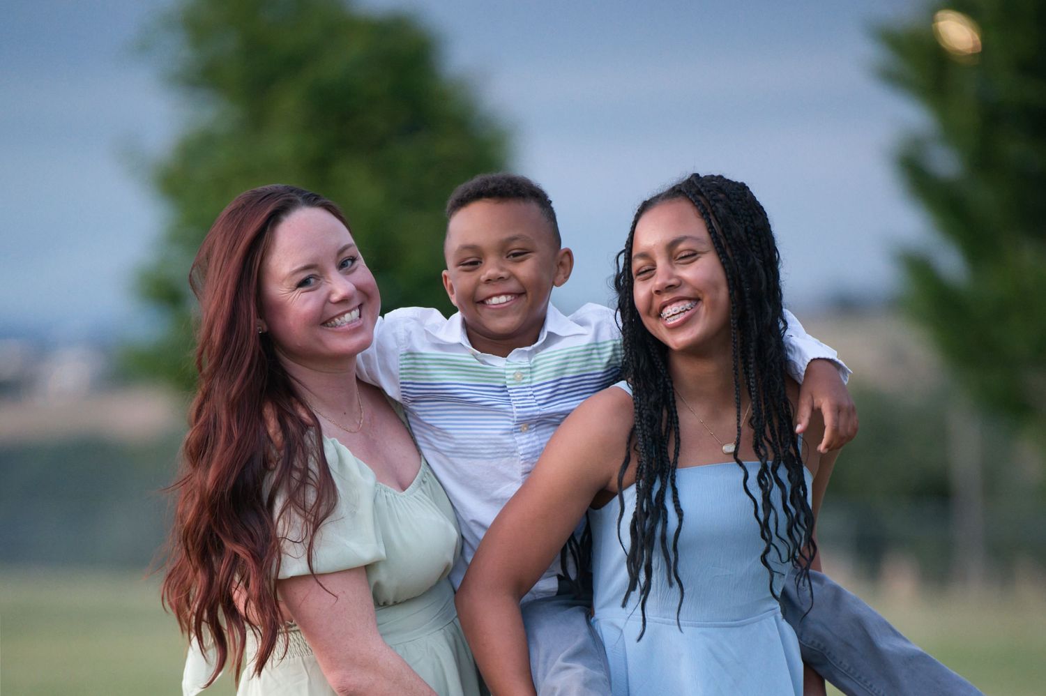 Two women hugging a young boy between them smiling outdoors in a green park at dusk.