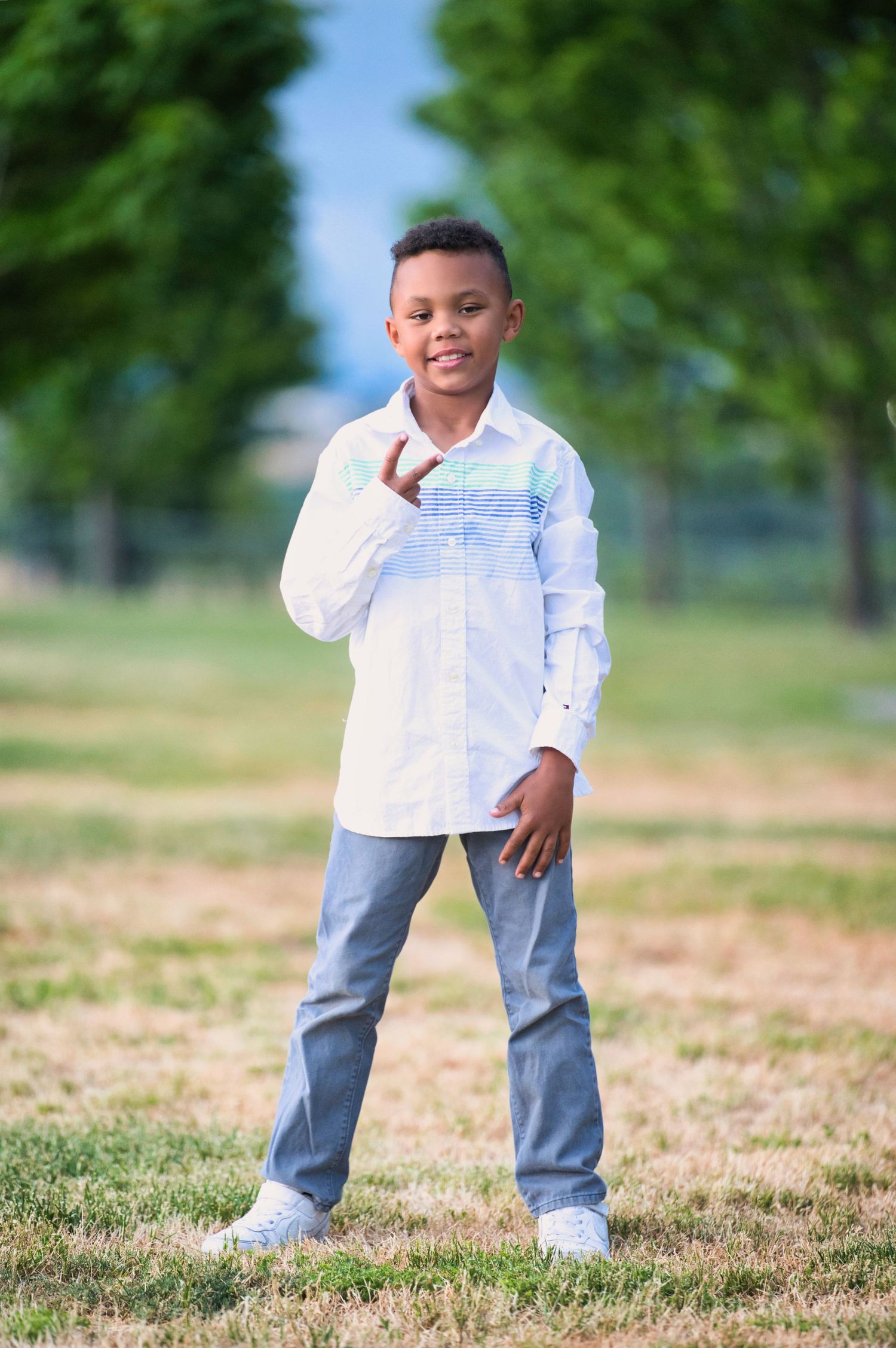 A young Black boy in a white button-up shirt and jeans poses confidently outdoors in a green park.
