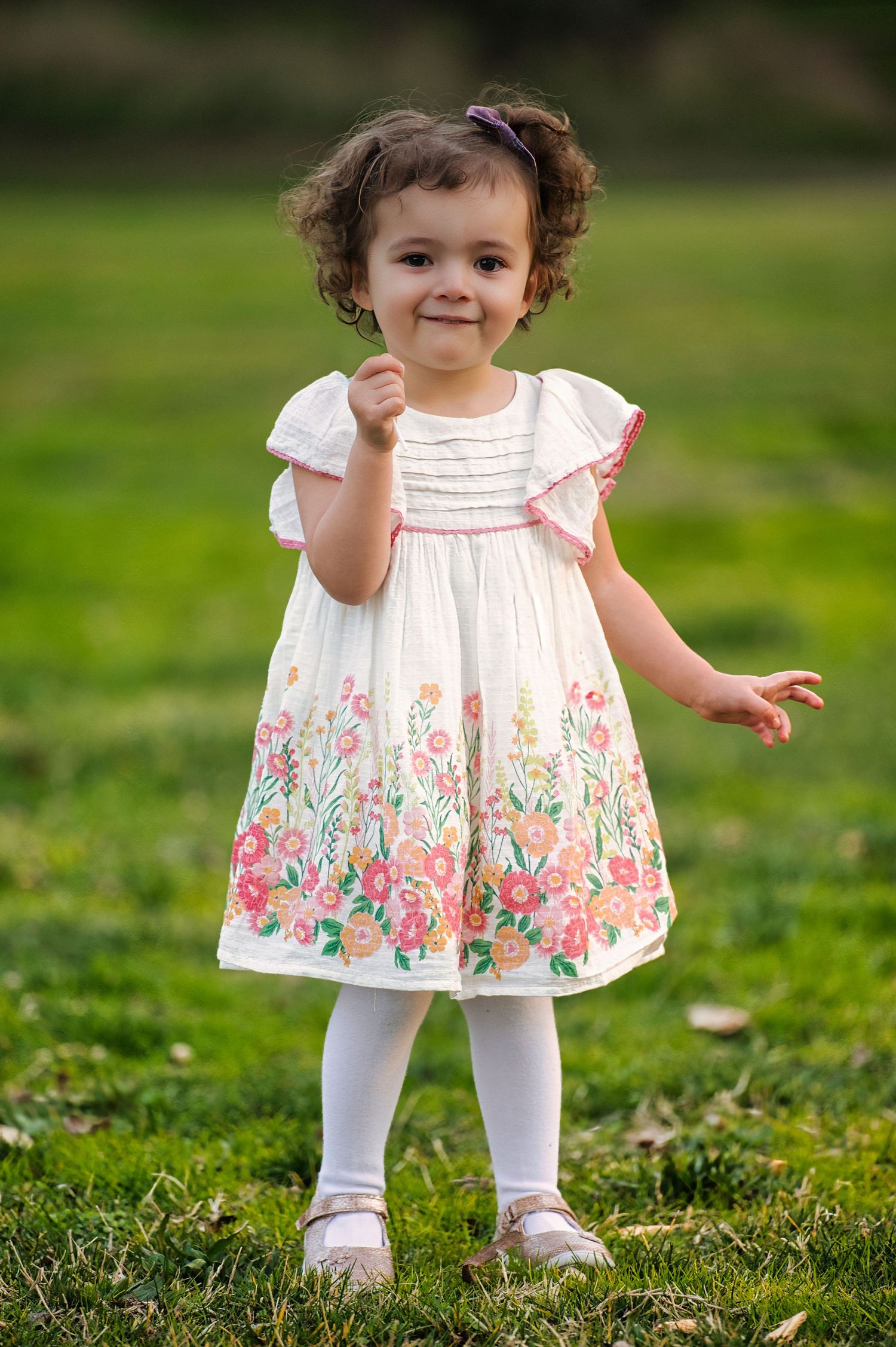Adorable toddler girl in a white floral dress with pink flowers standing on green grass outdoors.
