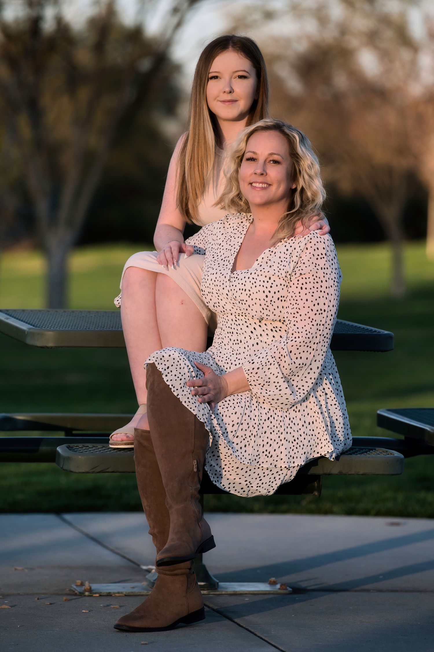 Woman in polka dot dress sits on bench with young girl standing behind her in a sunny park setting.