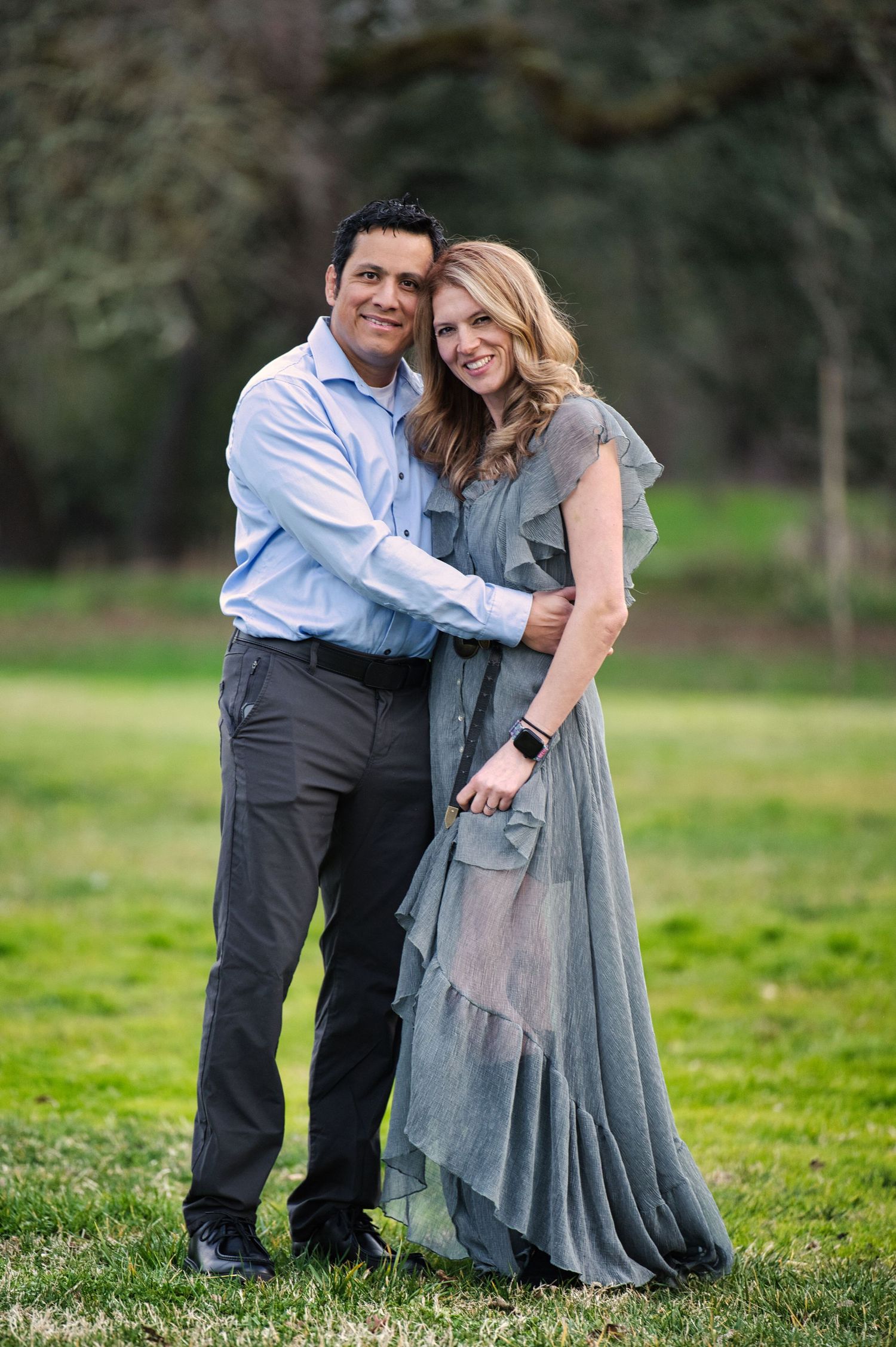 Couple embracing outdoors on green grass, man in light blue shirt, woman in elegant grey ruffle maxi dress.