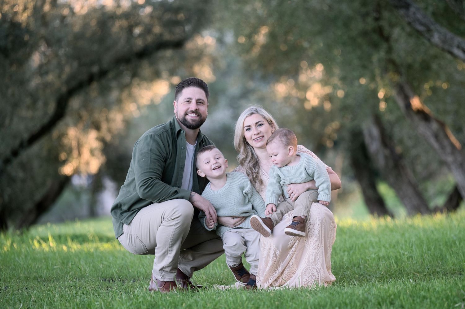 Happy family of four with two young boys smiling together on green grass in a sunny park.