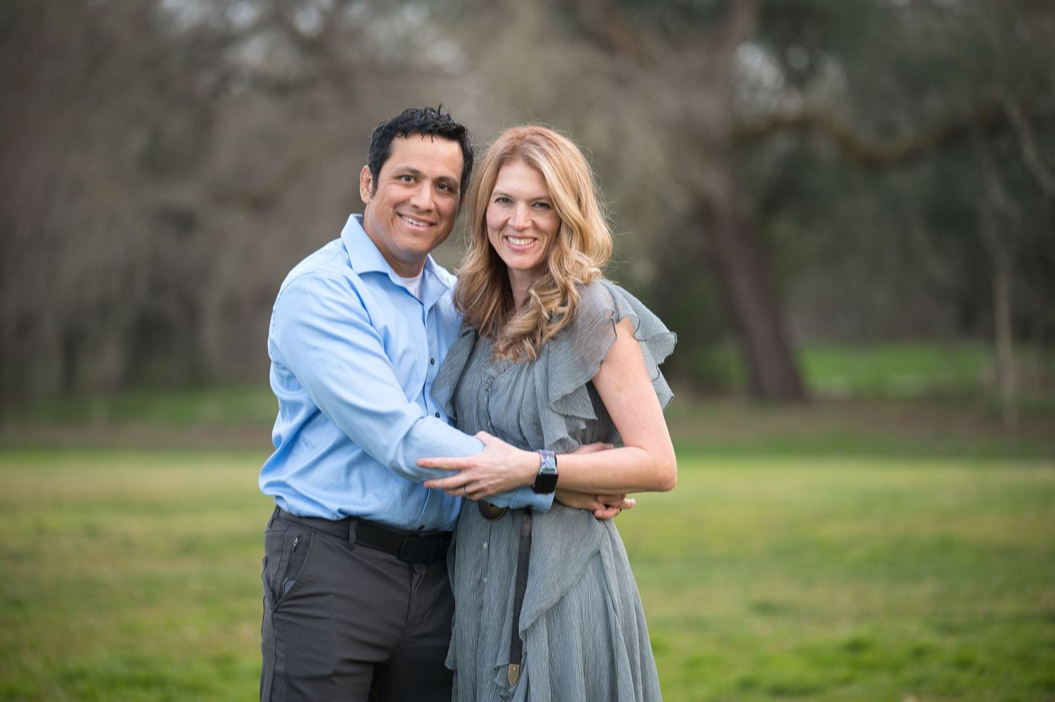 Smiling couple embracing in a green park, man in blue shirt and woman in grey ruffled dress.
