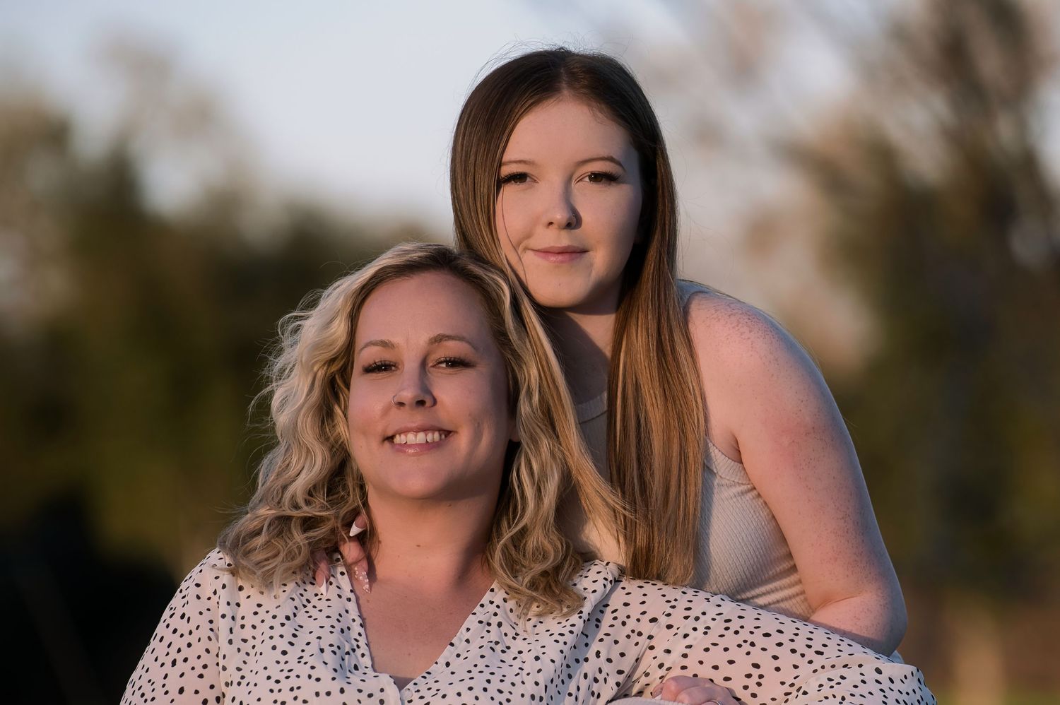 A smiling mother and teenage daughter pose closely together outdoors in warm golden light.