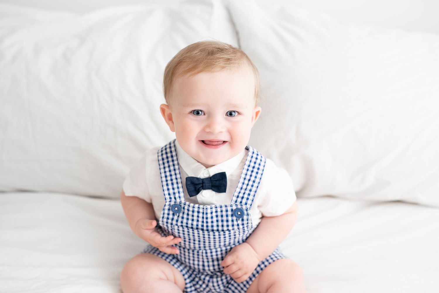 A small child wearing a navy gingham outfit with bow tie sits smiling on white bedding.