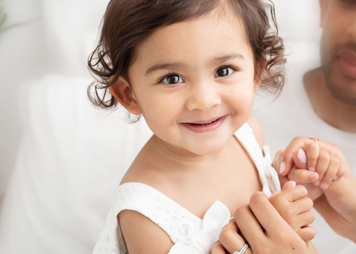 Smiling toddler in white dress with curly hair against light background.