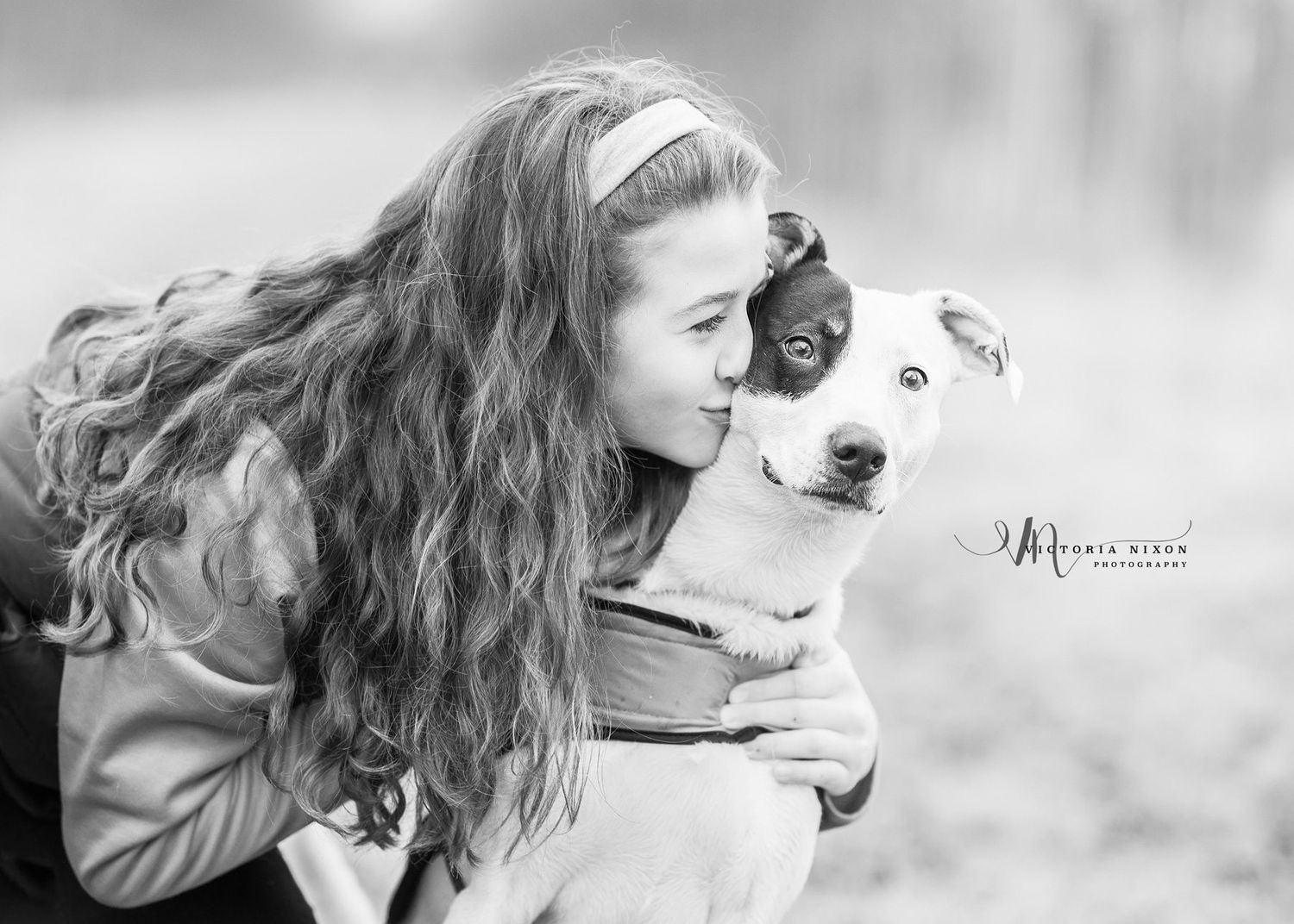 A black and white portrait shows a tender moment between a person with long wavy hair and their loving dog.