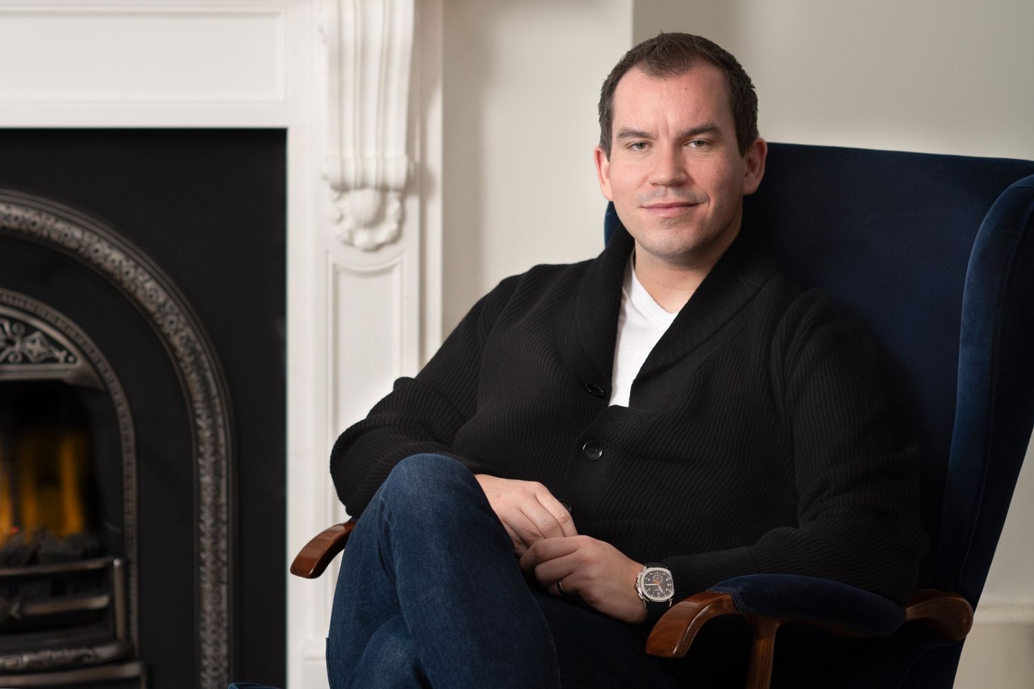 A person in a black sweater sits comfortably in a blue chair next to a traditional fireplace.