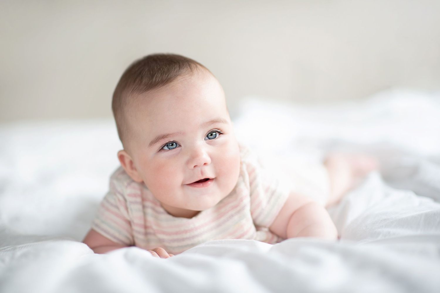Baby in striped outfit smiling while lying on white bedding.