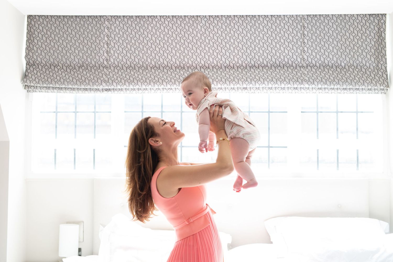 A mother in a coral top plays with her baby in a bright white room.