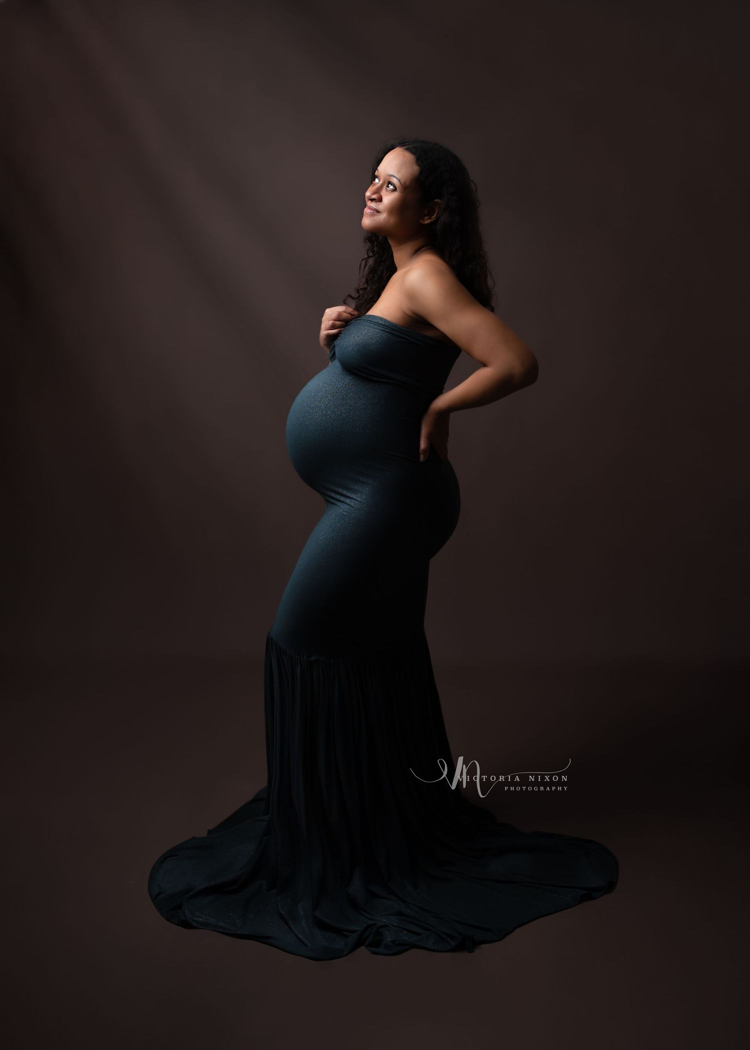 Dramatic maternity portraits in navy dress against dark background showcase elegant poses.