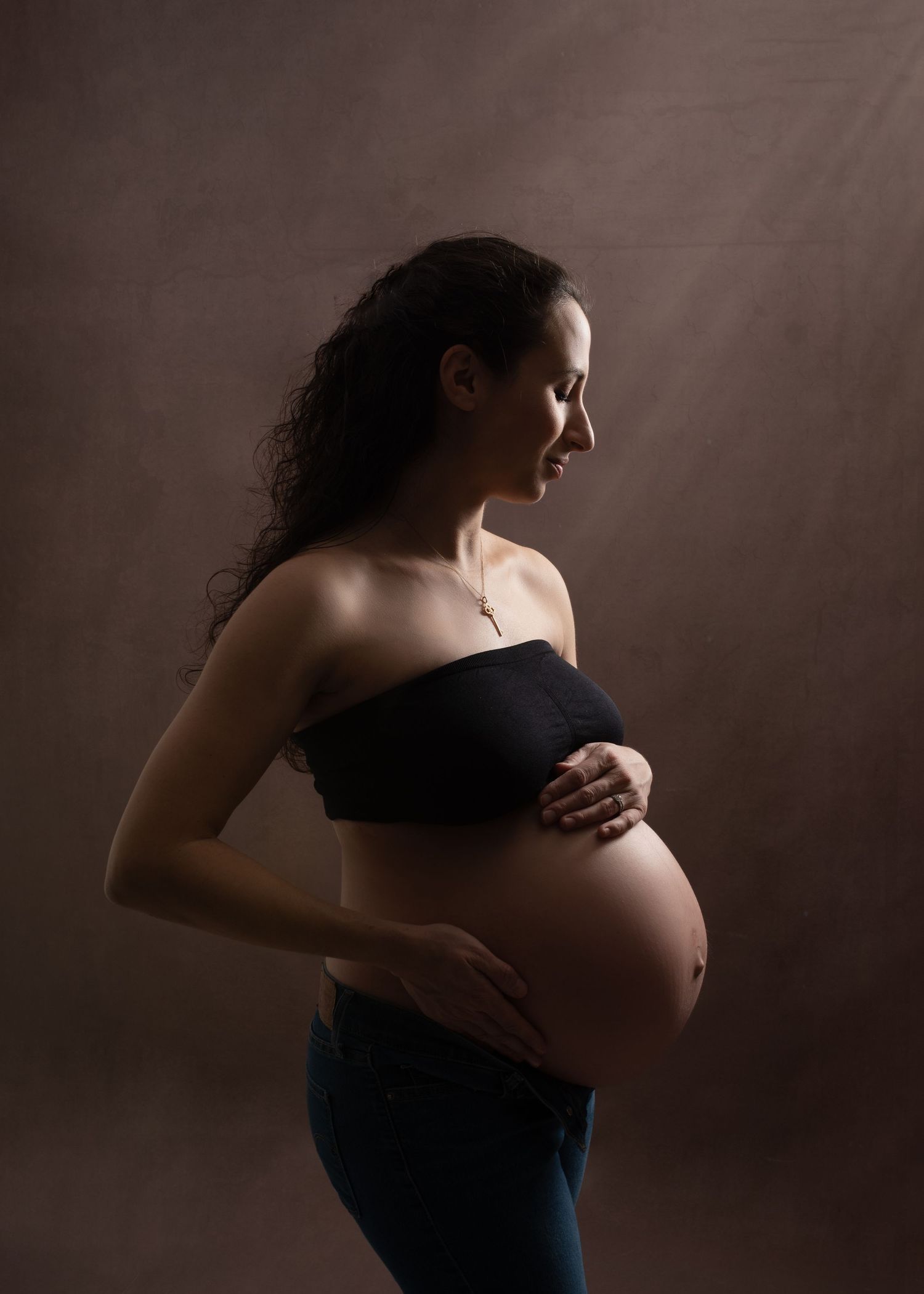 Silhouetted figure in dark dress stands against moody background in artistic maternity photo series.