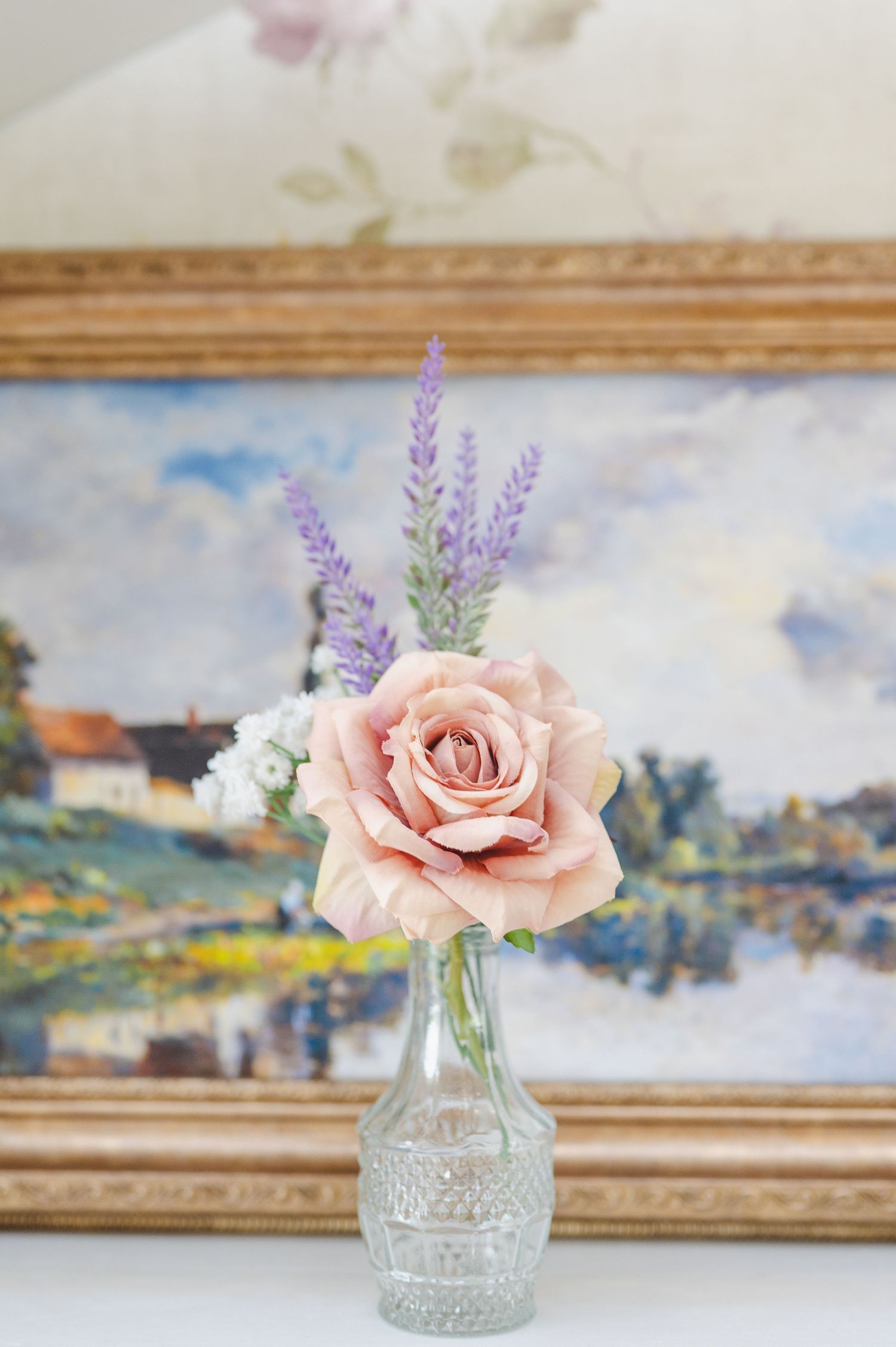 Delicate pink roses with lavender sprigs arranged in crystal vases against a soft vintage framed artwork background.