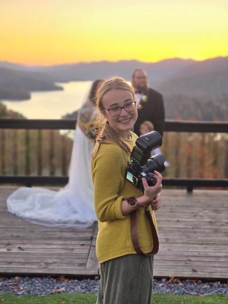 A photographer in a yellow sweater holds a camera on a deck overlooking a lake and mountains at sunset at The Blue Montanya Resort in TN.