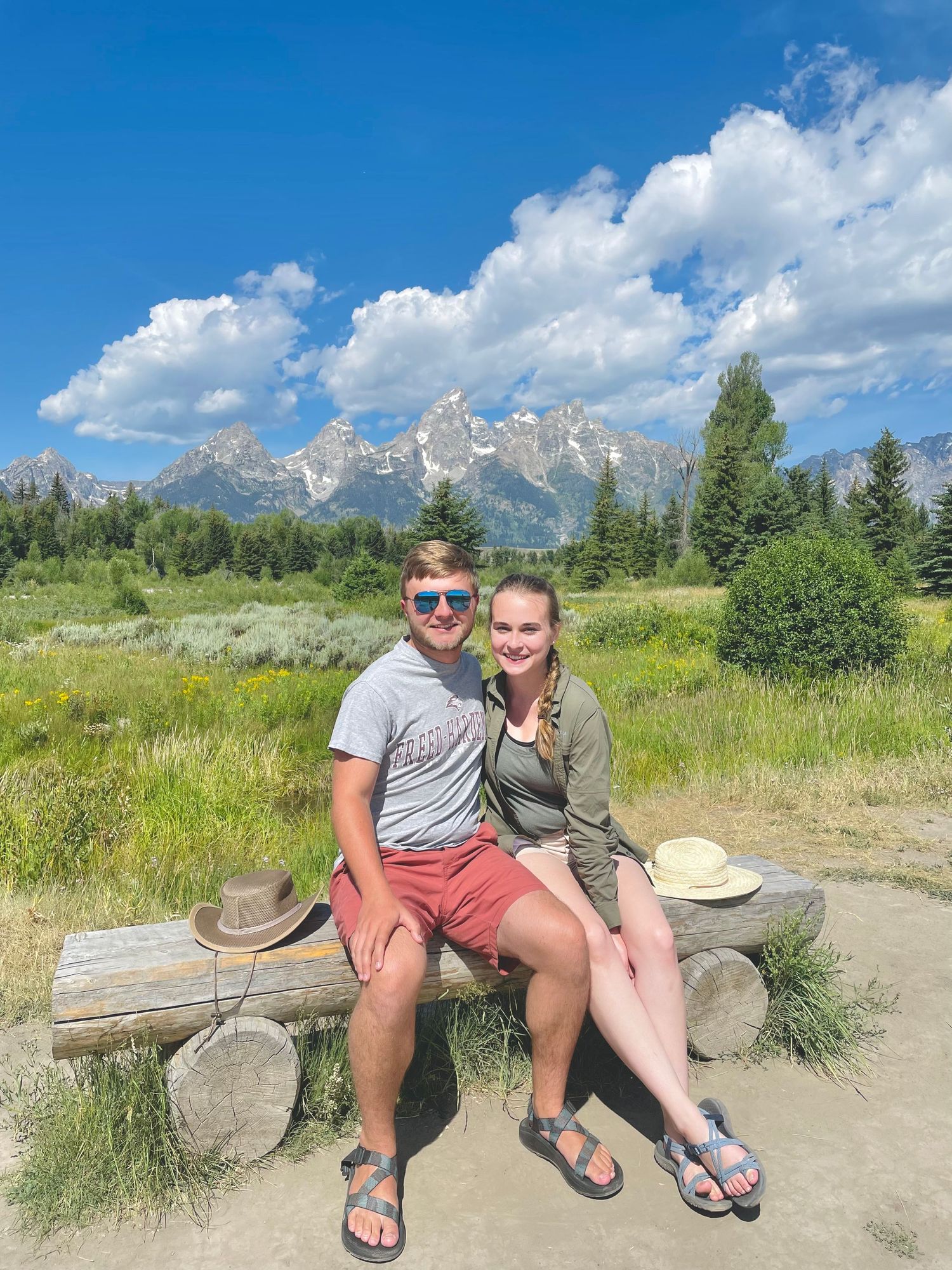 Two people enjoy a sunny summer day sitting on a wooden bench with the Grand Teton mountains rising majestically in the background.