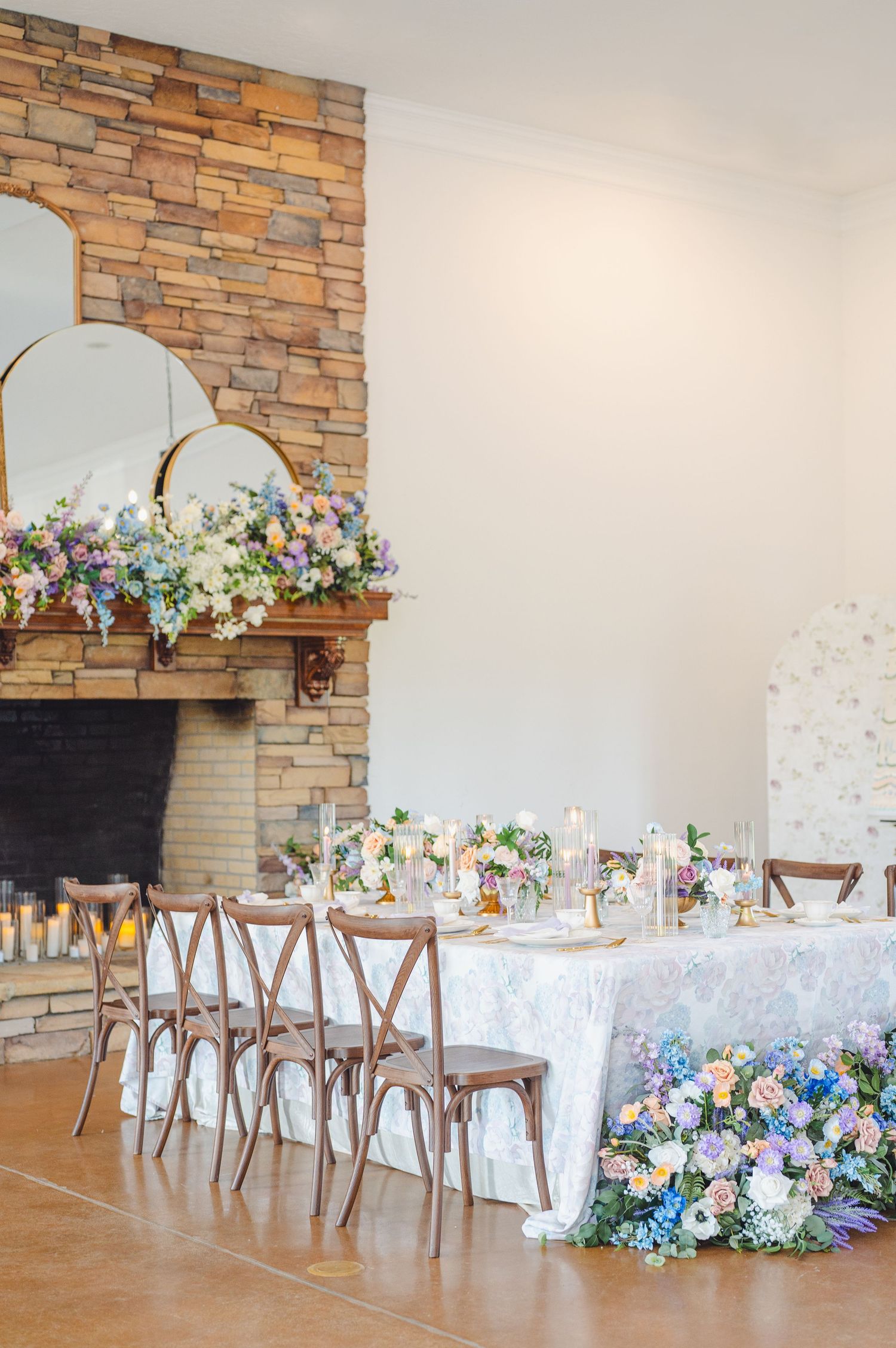 Elegant wedding reception table with wooden chairs against stone fireplace decorated with pastel floral arrangements.