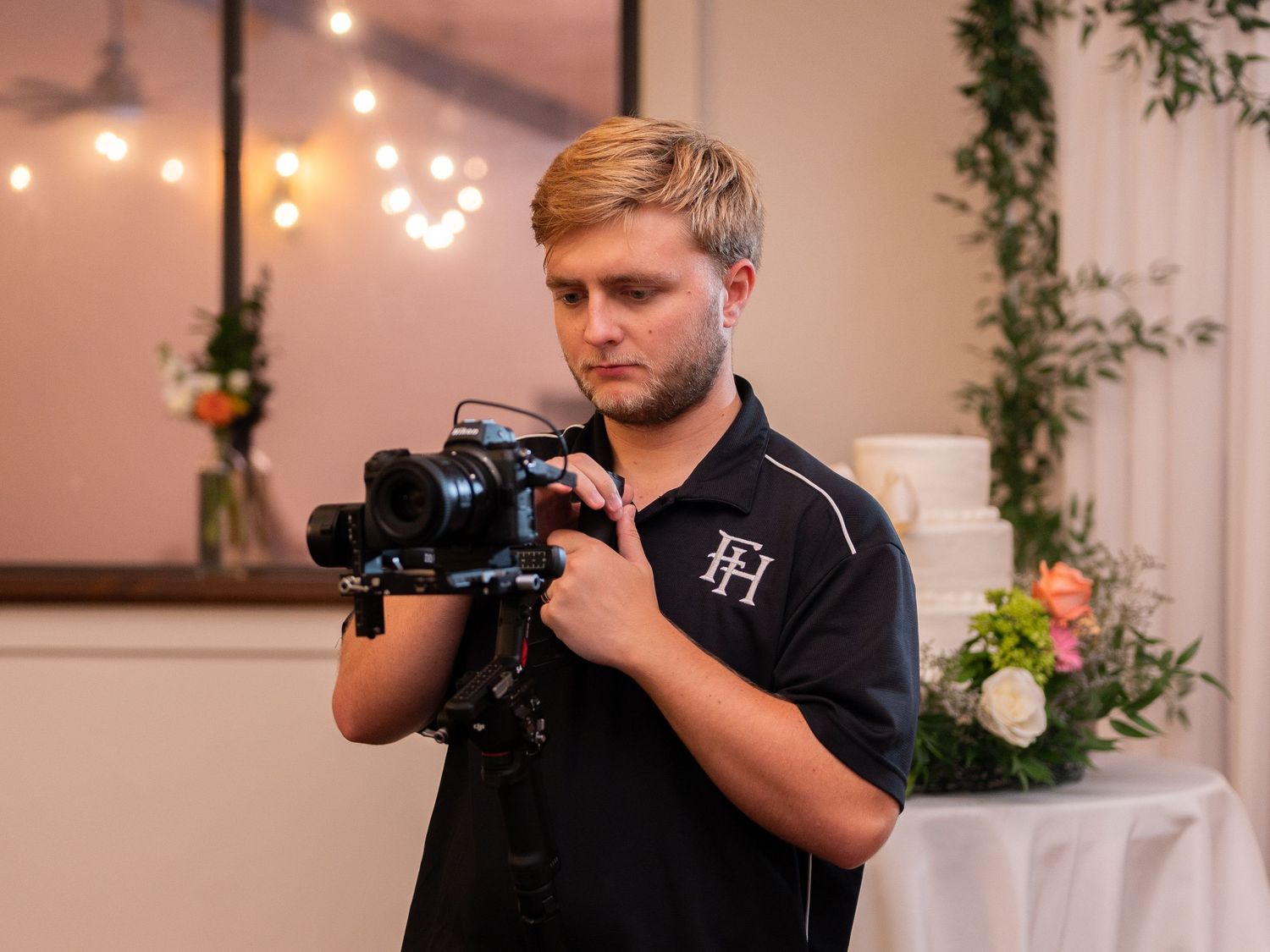 Videographer operating a professional camera at an event with string lights and floral decorations in the background.