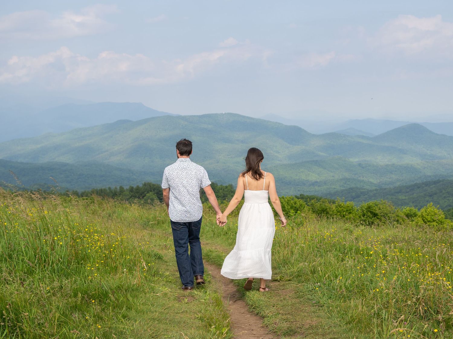 A couple holding hands walks along a mountain trail surrounded by rolling green hills under a light blue sky.
