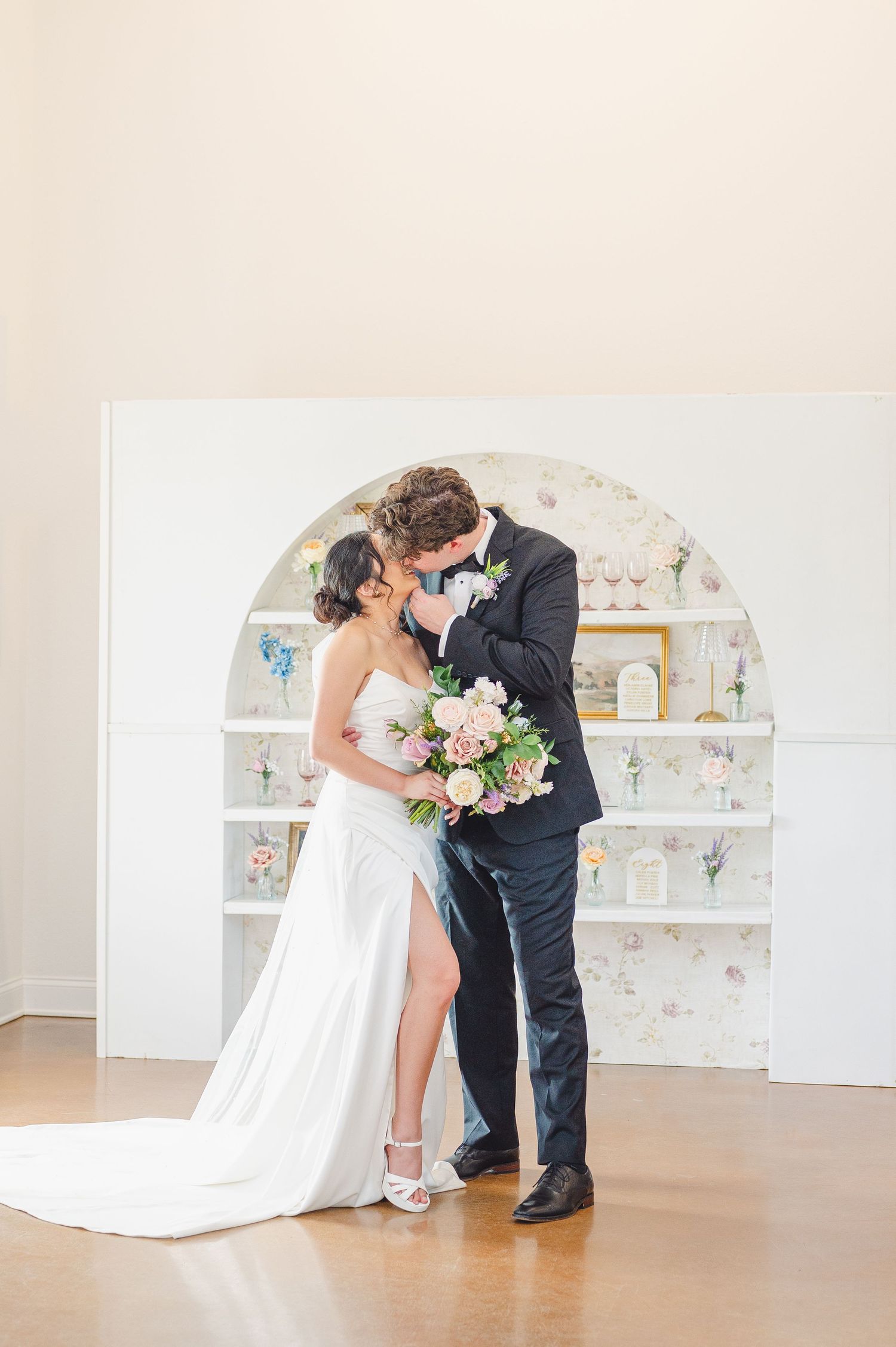 A couple shares a romantic moment in front of a vintage-style arched display shelf, with pink and white floral bouquet.