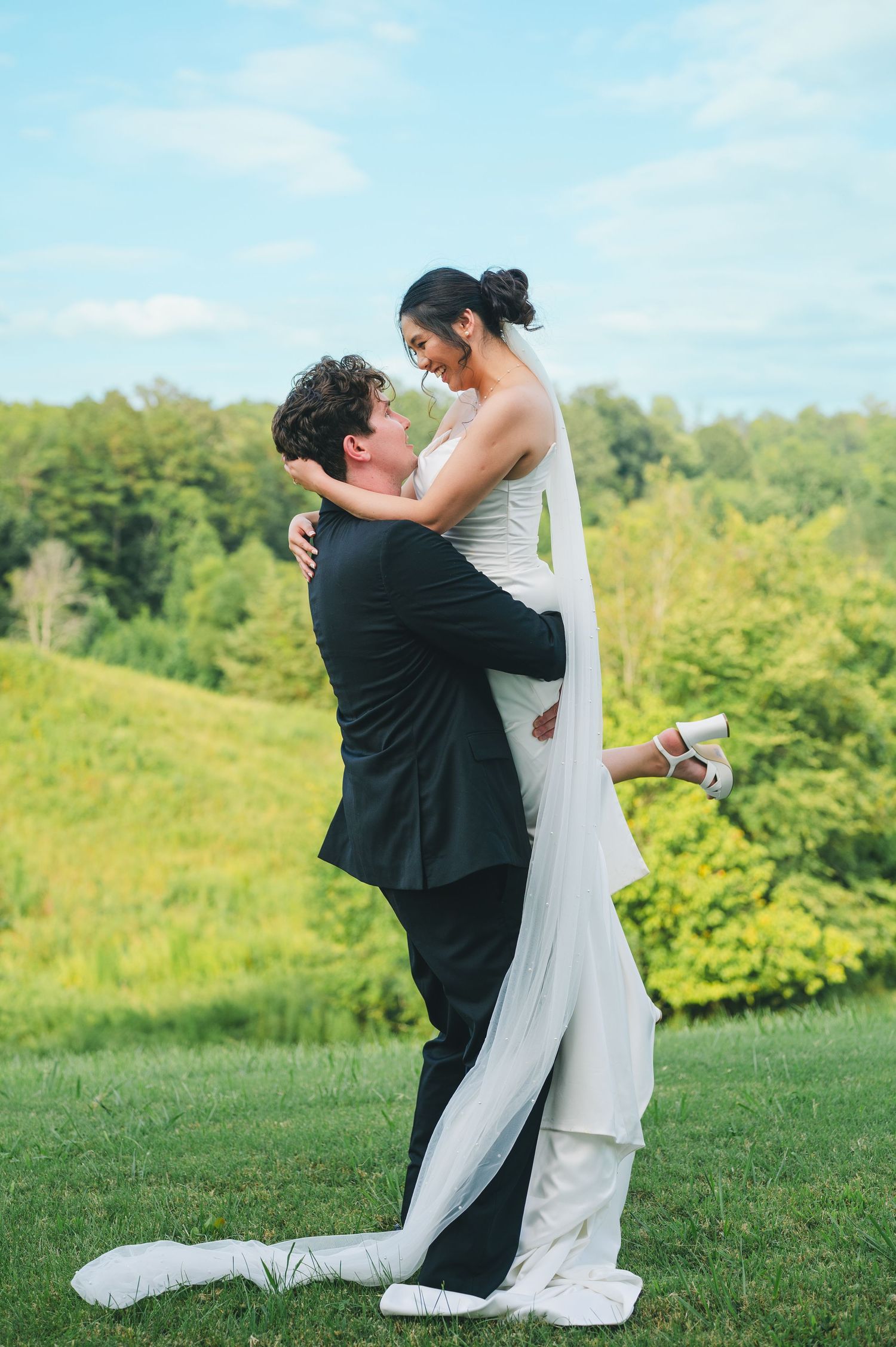 A couple embraces on a grassy hilltop during their wedding celebration with green trees in the background.