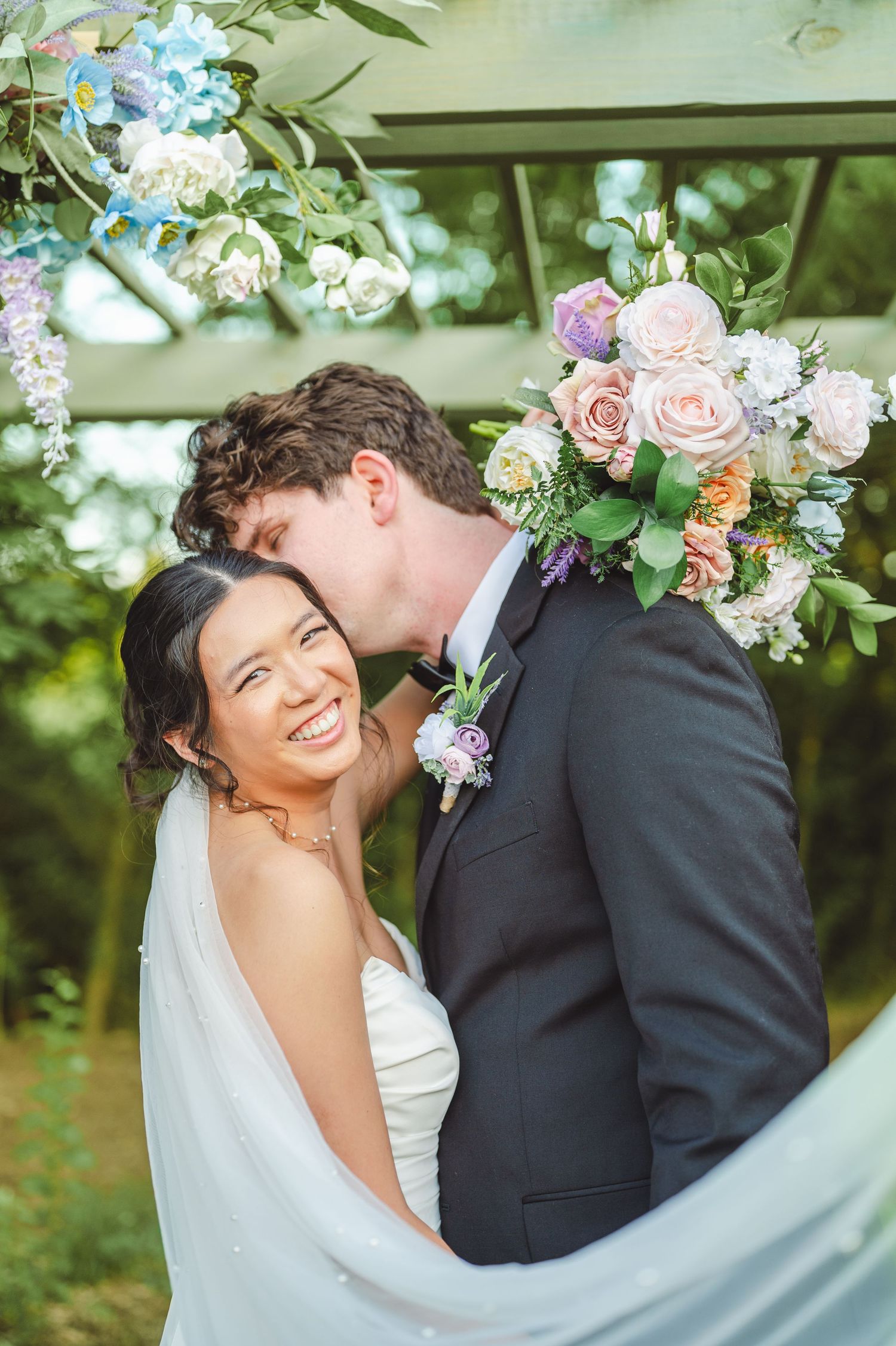 Wedding couple sharing an intimate moment under floral arch with pastel roses and greenery decorations.