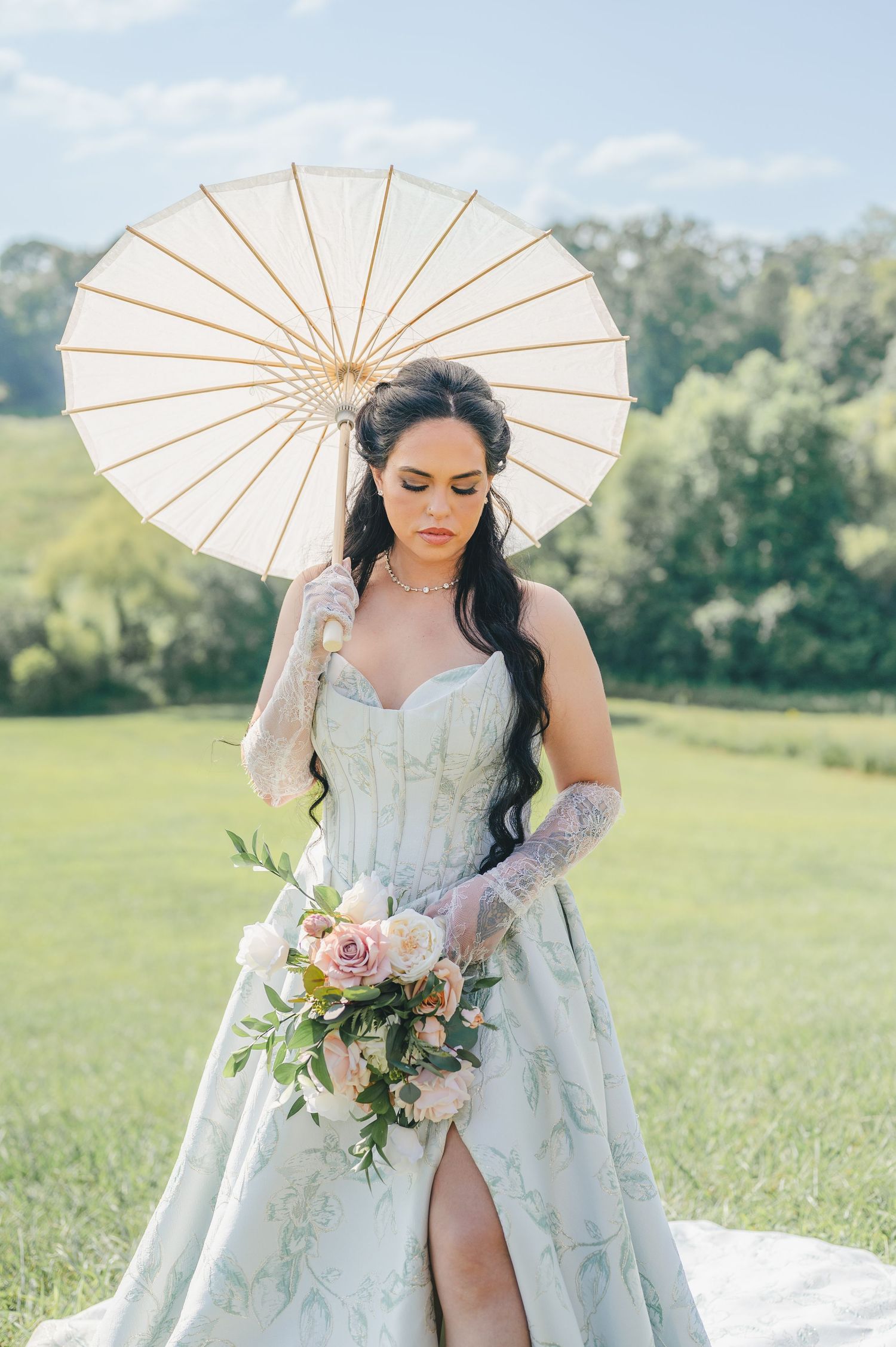 A bride in a floral blue wedding gown holds a parasol and bouquet of pink flowers in a sunny meadow.