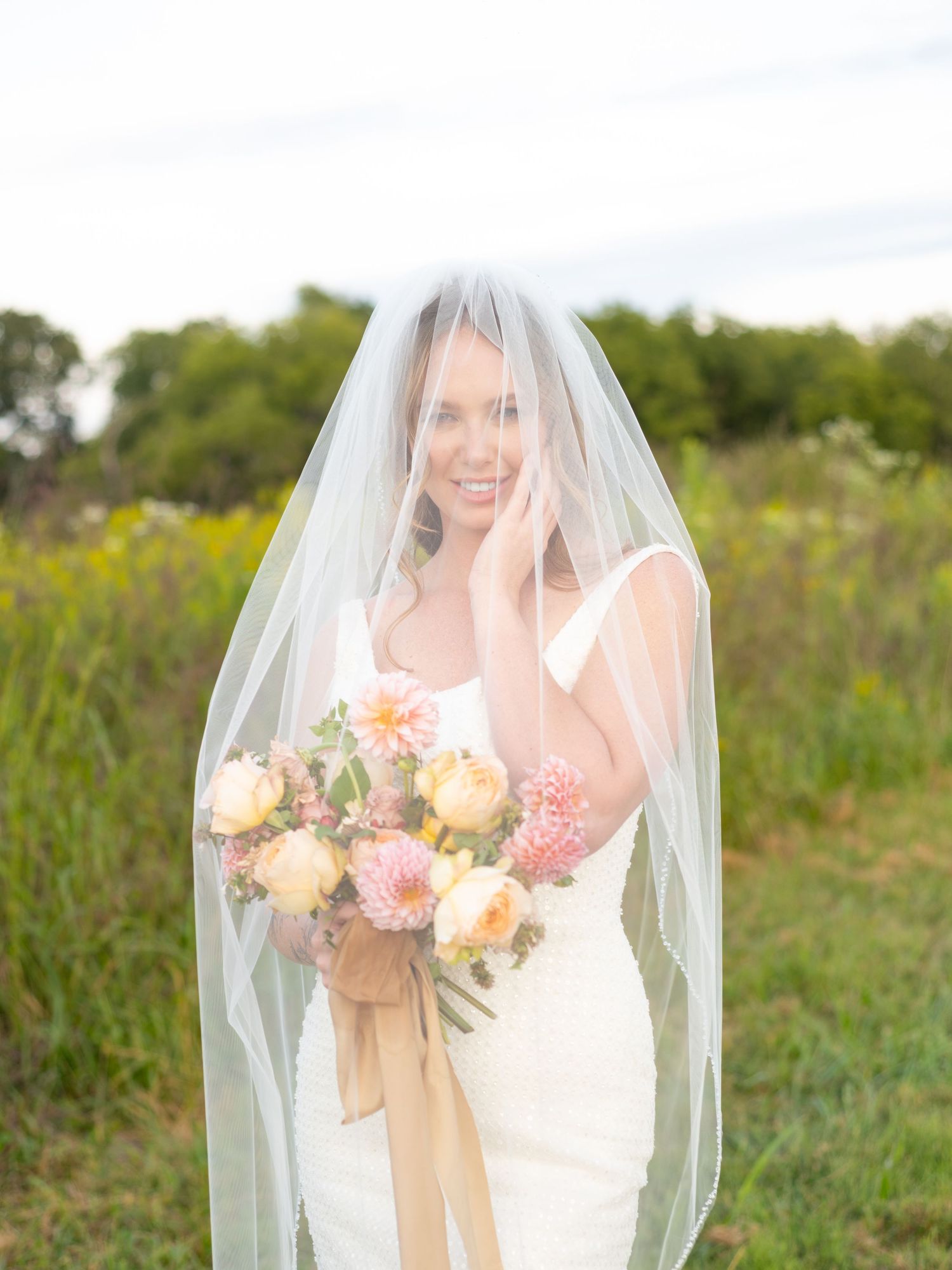 A bride in a white dress and veil holds a beautiful bouquet of peach roses and pink dahlias in a sunlit meadow.