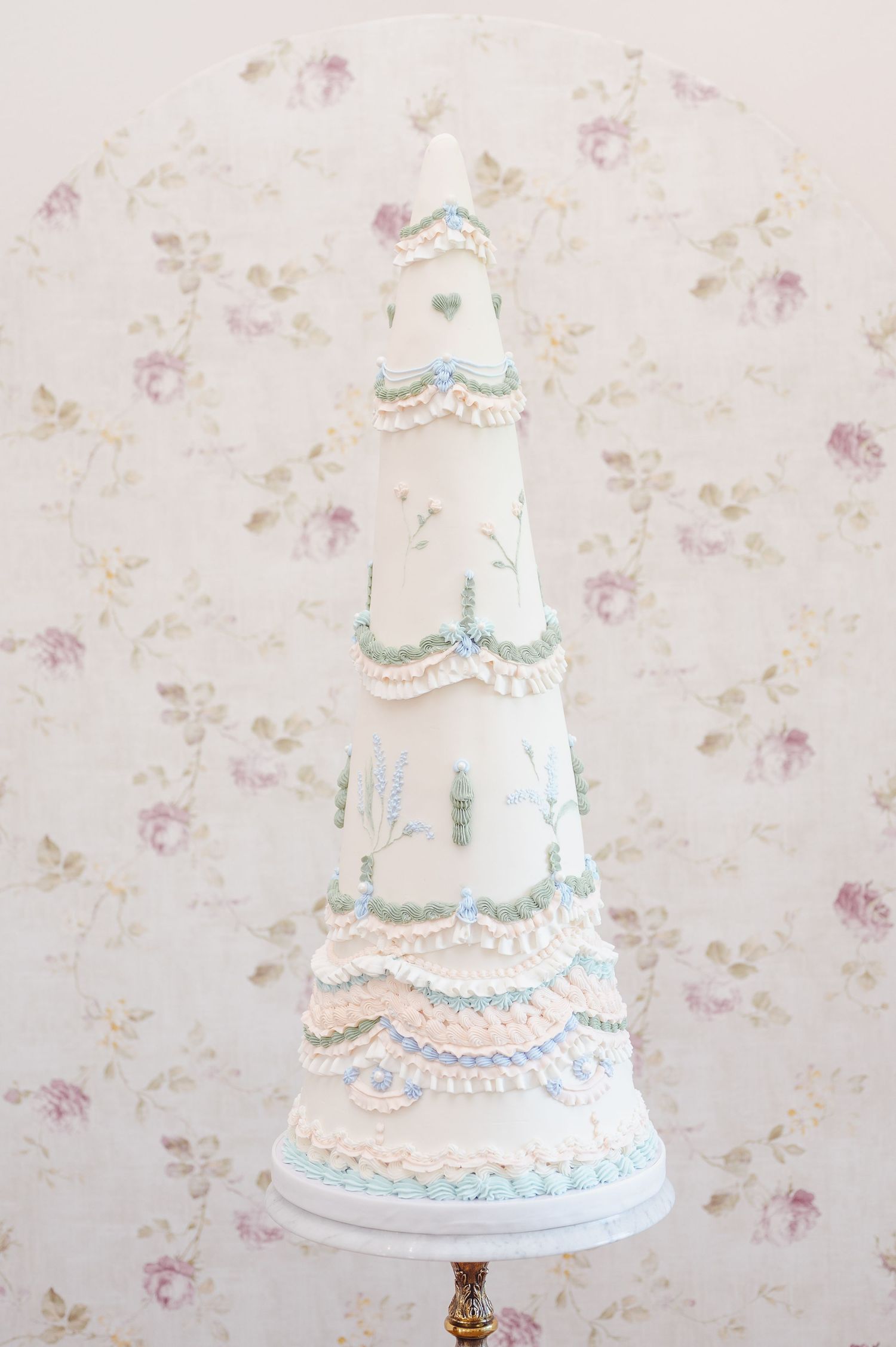 A series of wedding cakes with delicate lace-like piping and peach accents displayed against a floral wallpaper background.