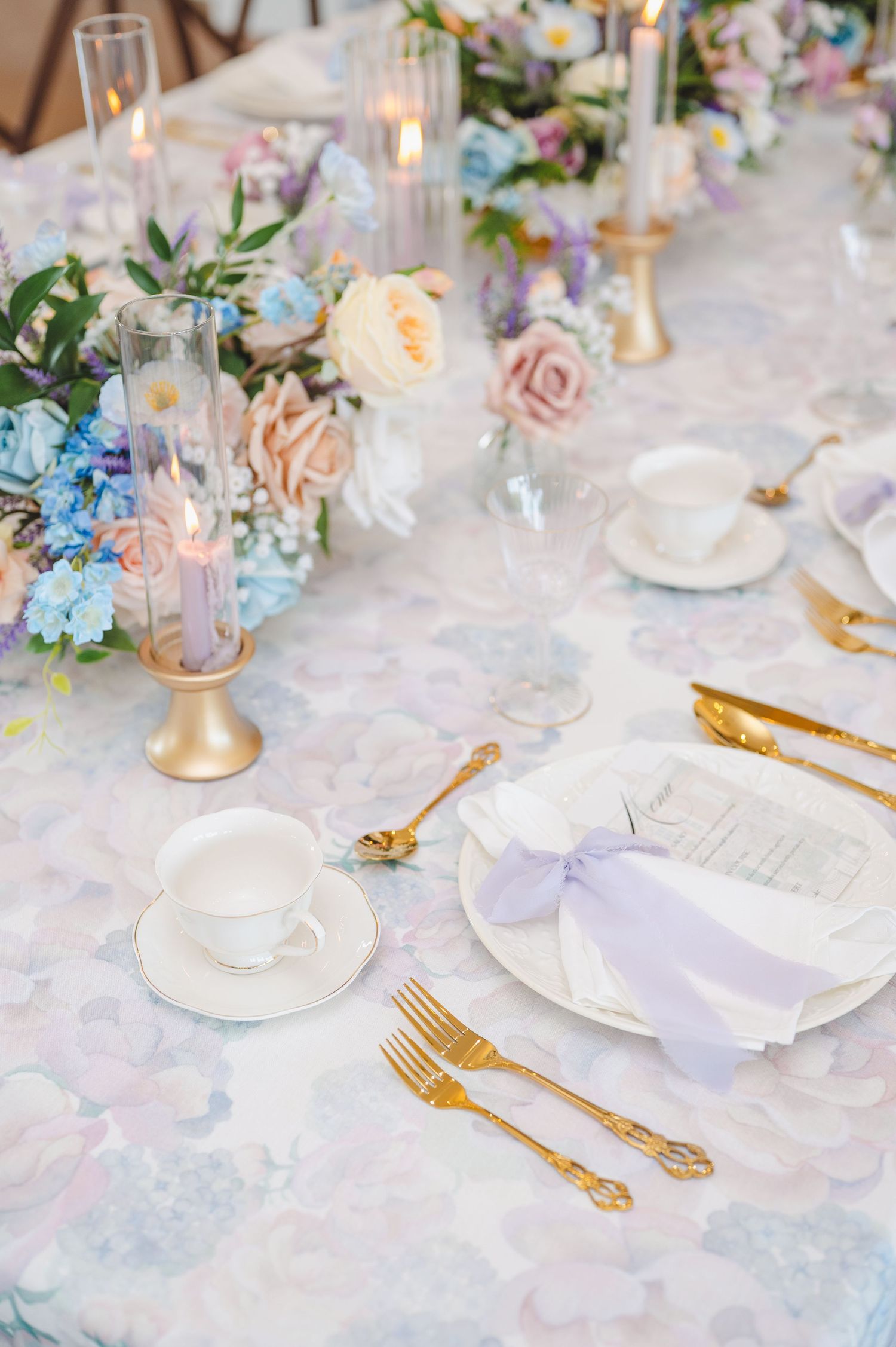 Elegant wedding table setting with pastel blue and pink flowers, gold candlesticks, and fine china on a delicate floral tablecloth.