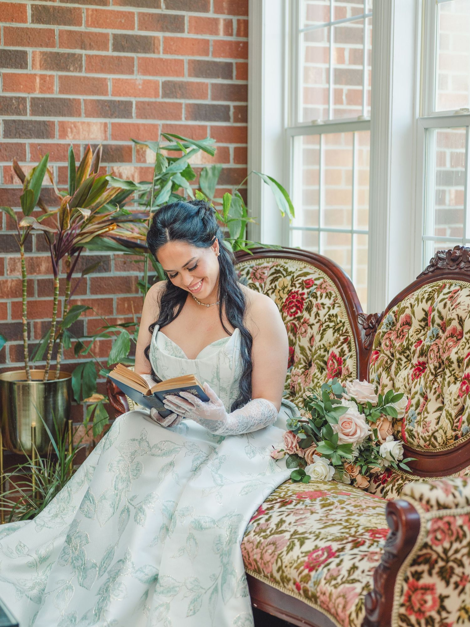A bride in a white wedding gown relaxes on an antique floral-patterned chaise lounge against a brick wall with plants.