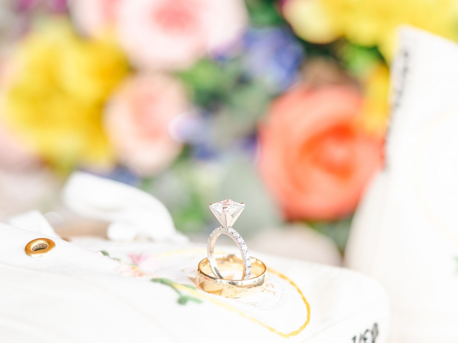 Wedding rings and a diamond engagement ring displayed with soft pastel flowers in the background.