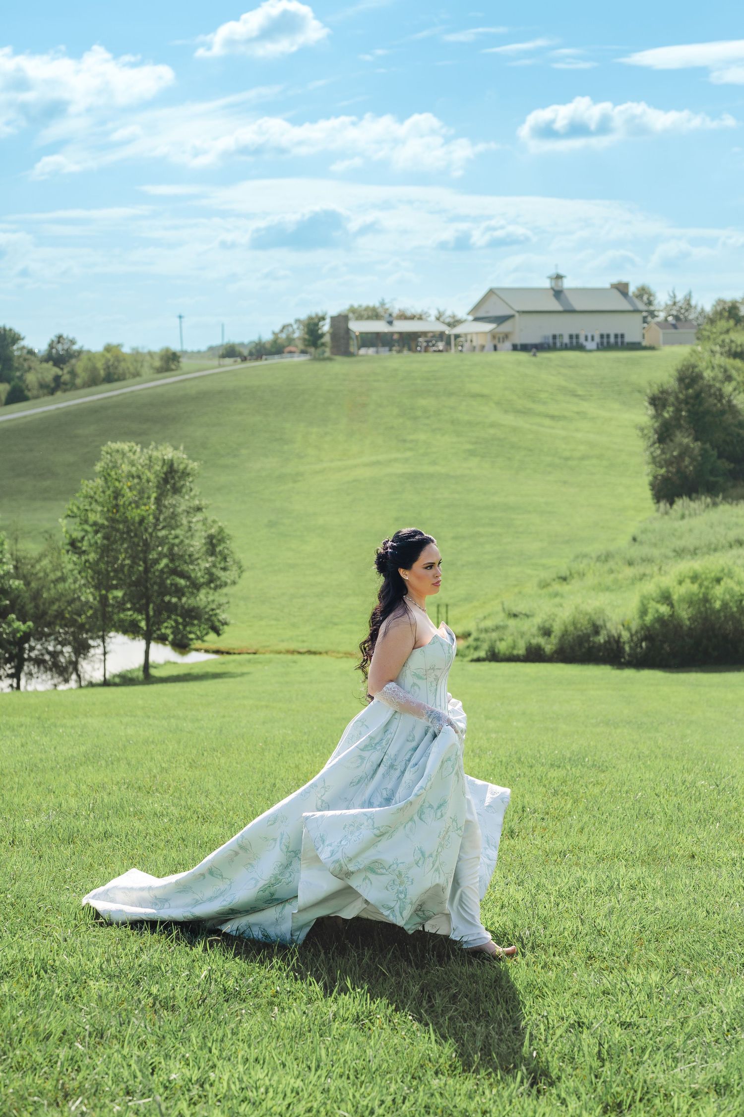 A person in a flowing light blue dress sits gracefully in a rolling green meadow on a sunny summer day.