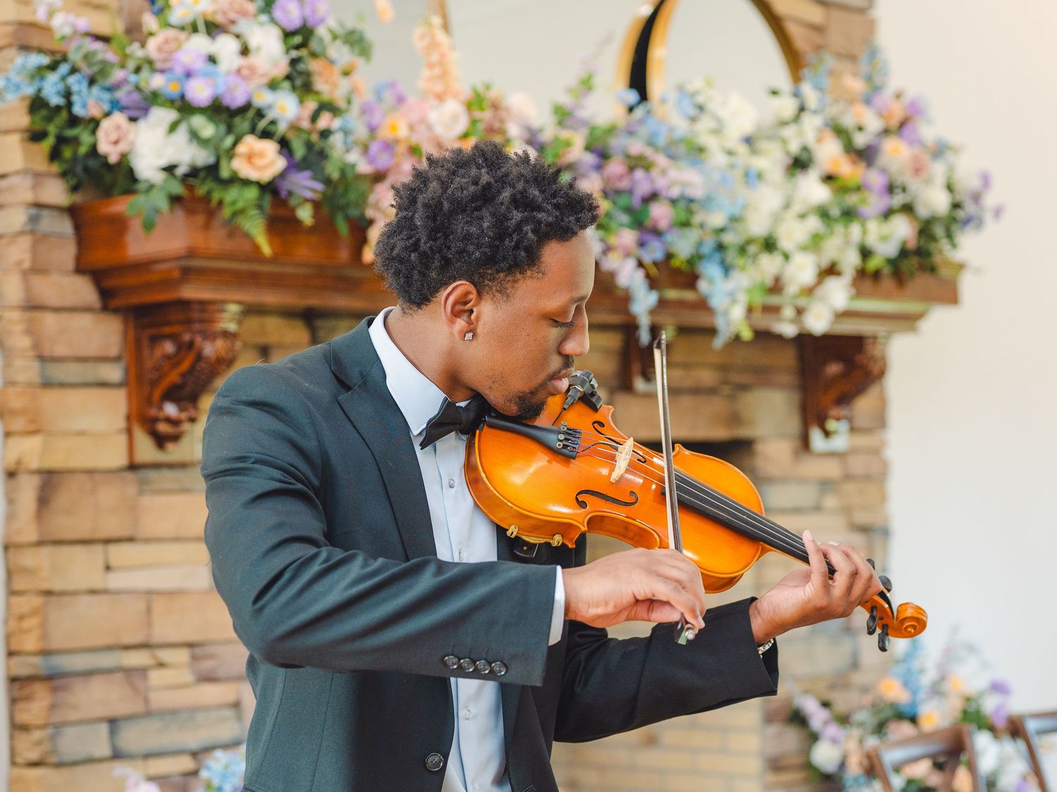 A violinist performs in formal attire against a backdrop of floral arrangements and stone archway at an event.