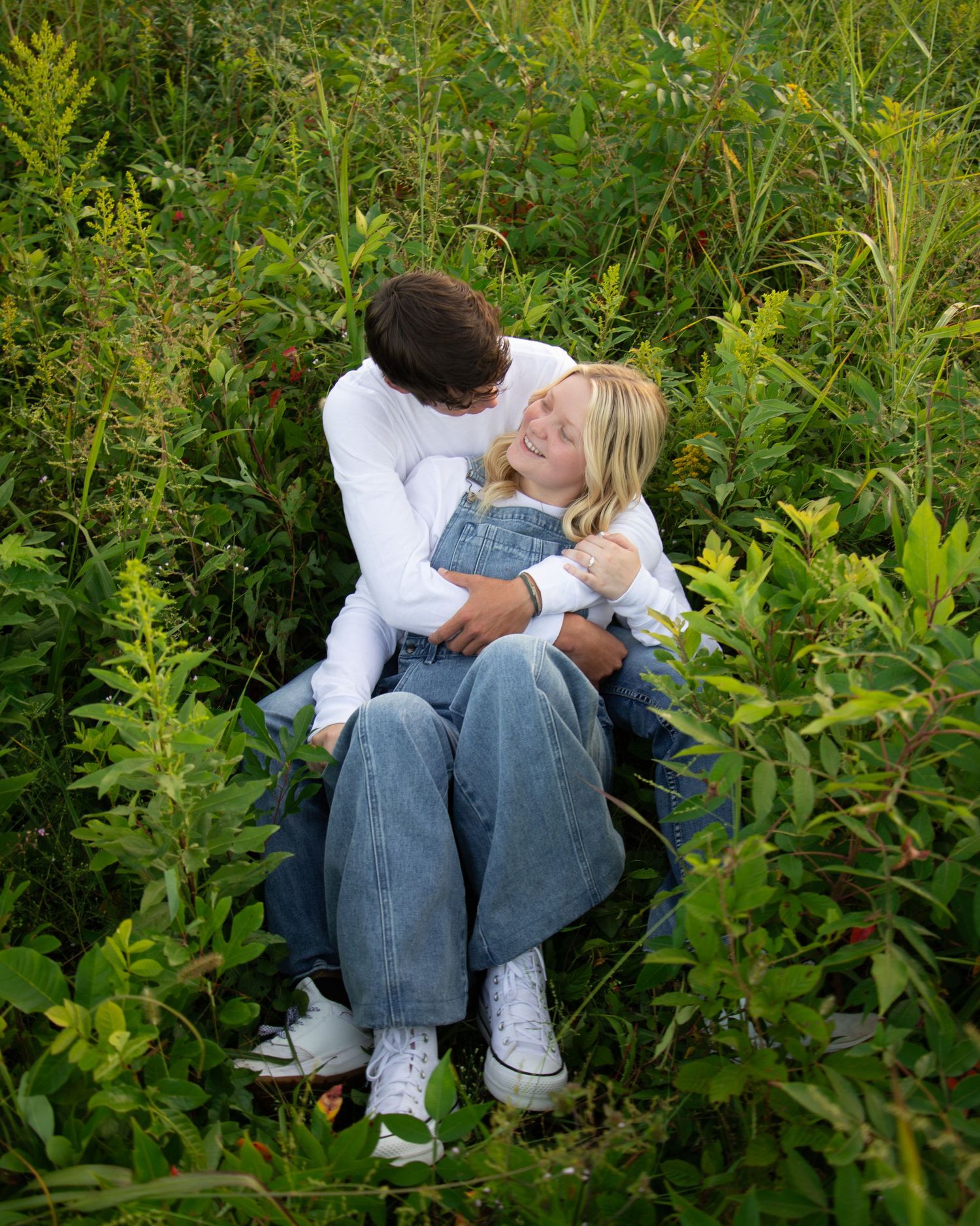 A tender embrace between two people sitting among lush green foliage on a summer day.