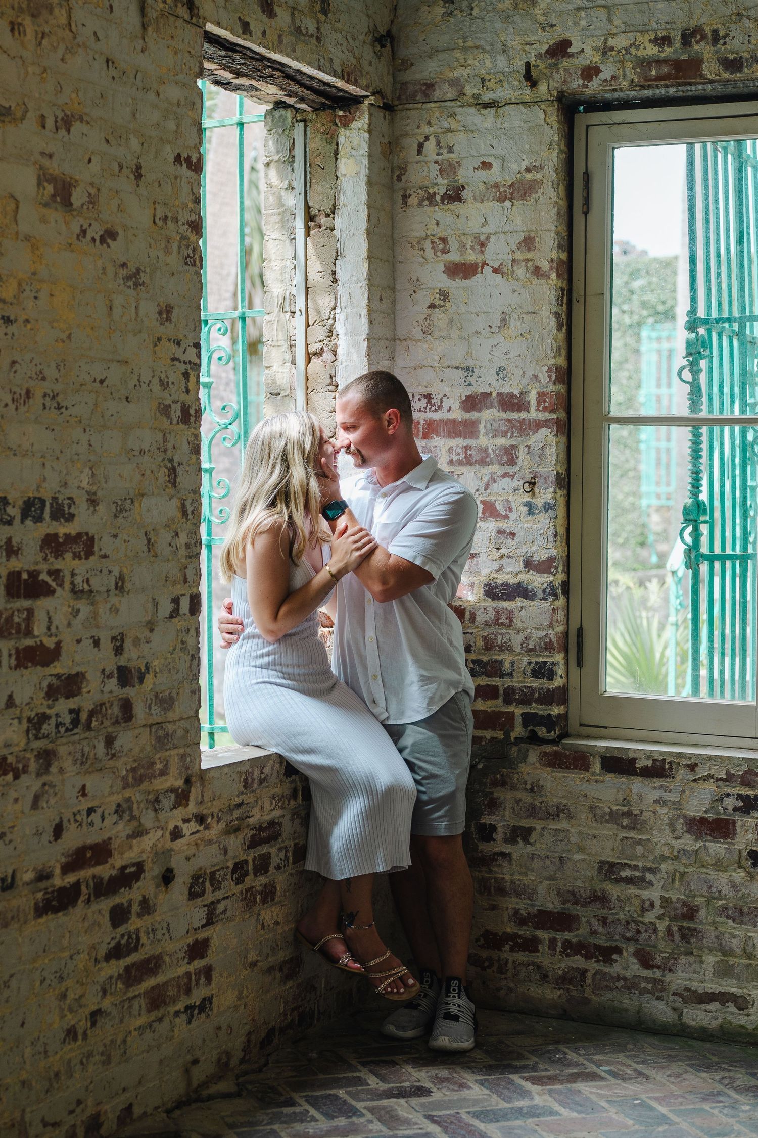 A romantic couple embraces by a rustic brick window with teal shutters in an old industrial building setting.