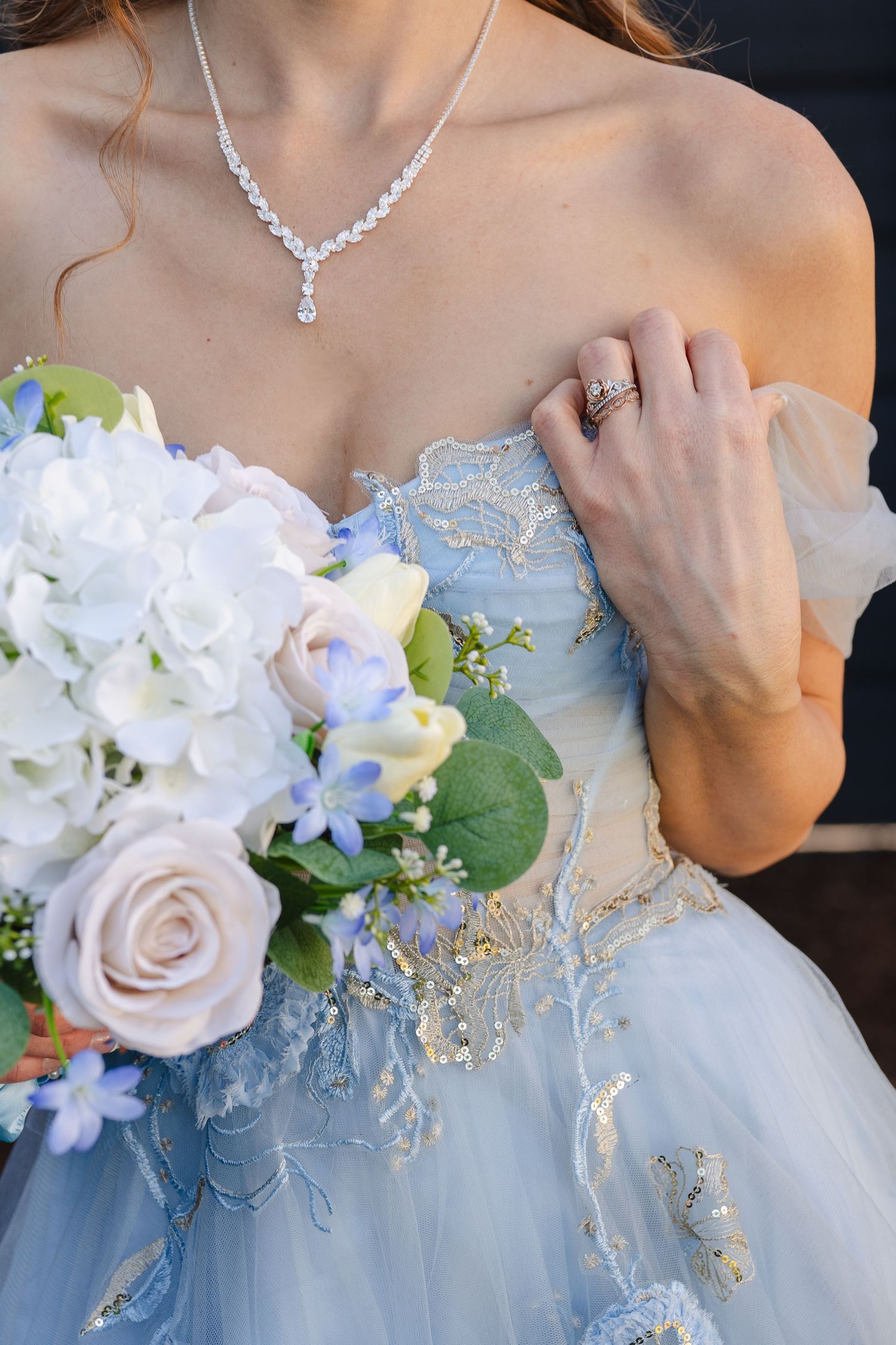 Close-up of a blue lace wedding gown with delicate crystal necklace and pastel rose and delphinium bouquet.