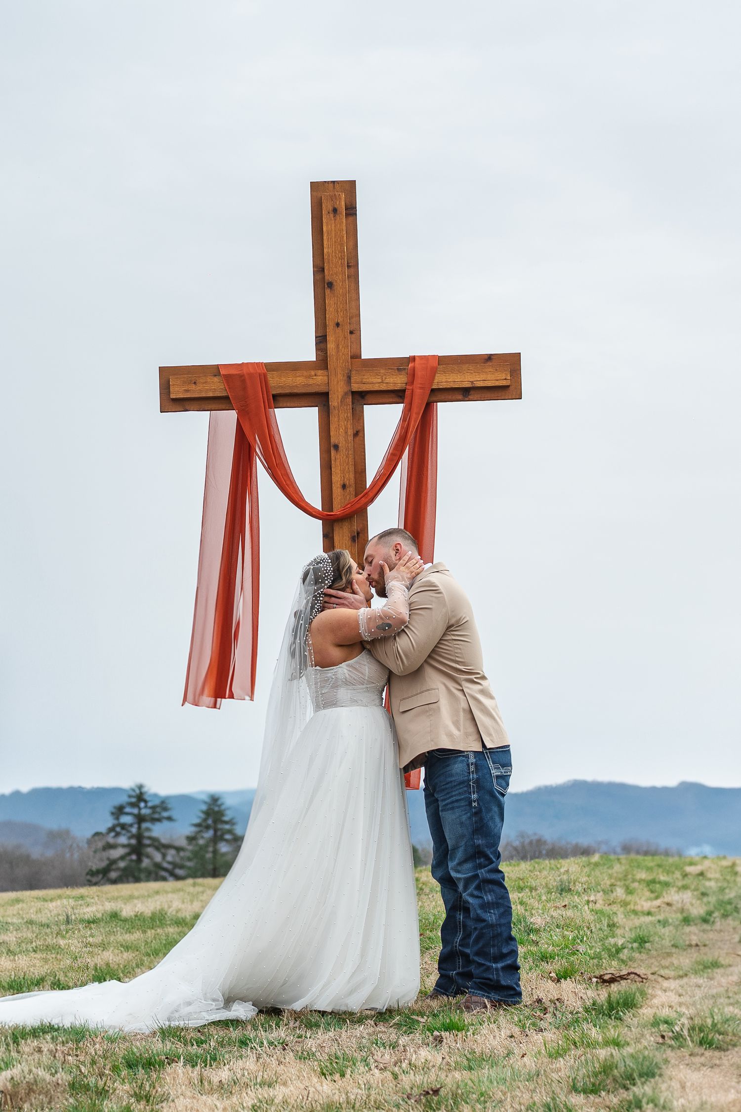 A couple shares their wedding moment in front of a wooden cross with red draping on a hilltop with North Carolina mountain views.