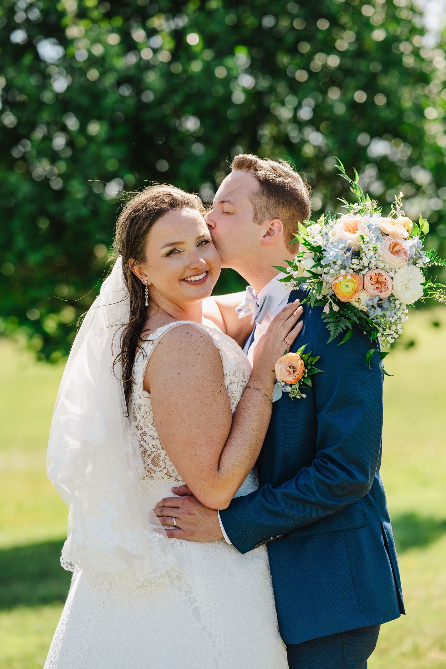Joyful wedding couple embraces outdoors with peach and white floral bouquet against green garden background.