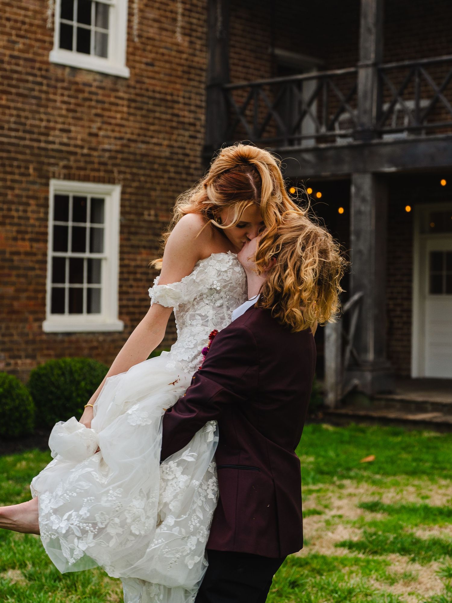 A couple shares a romantic moment in front of a brick building, with person in burgundy lifting someone in a white lace gown.