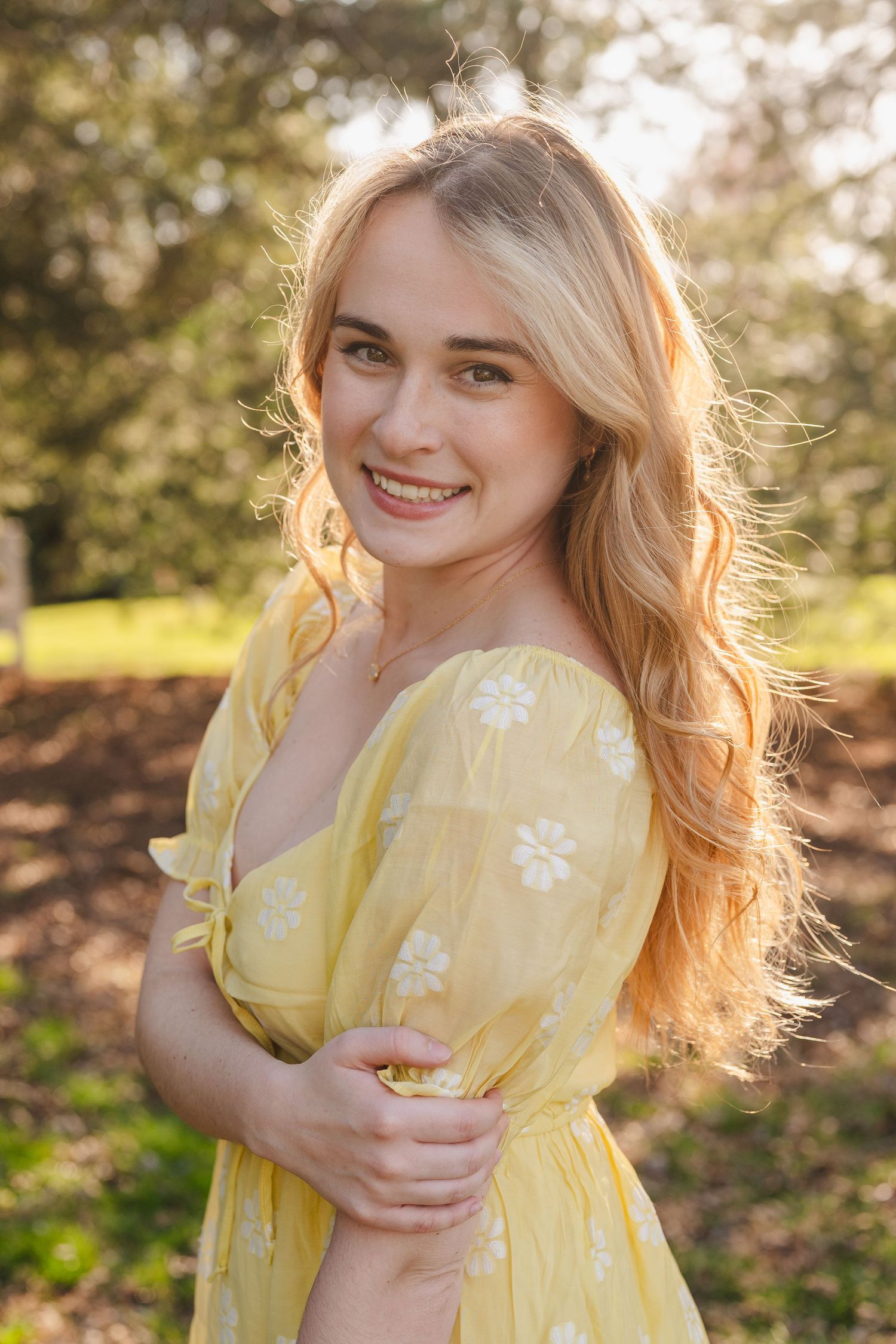 Sydney Johnson a wedding photographer in a yellow floral dress poses outdoors in warm sunlight with long blonde hair flowing in the breeze.