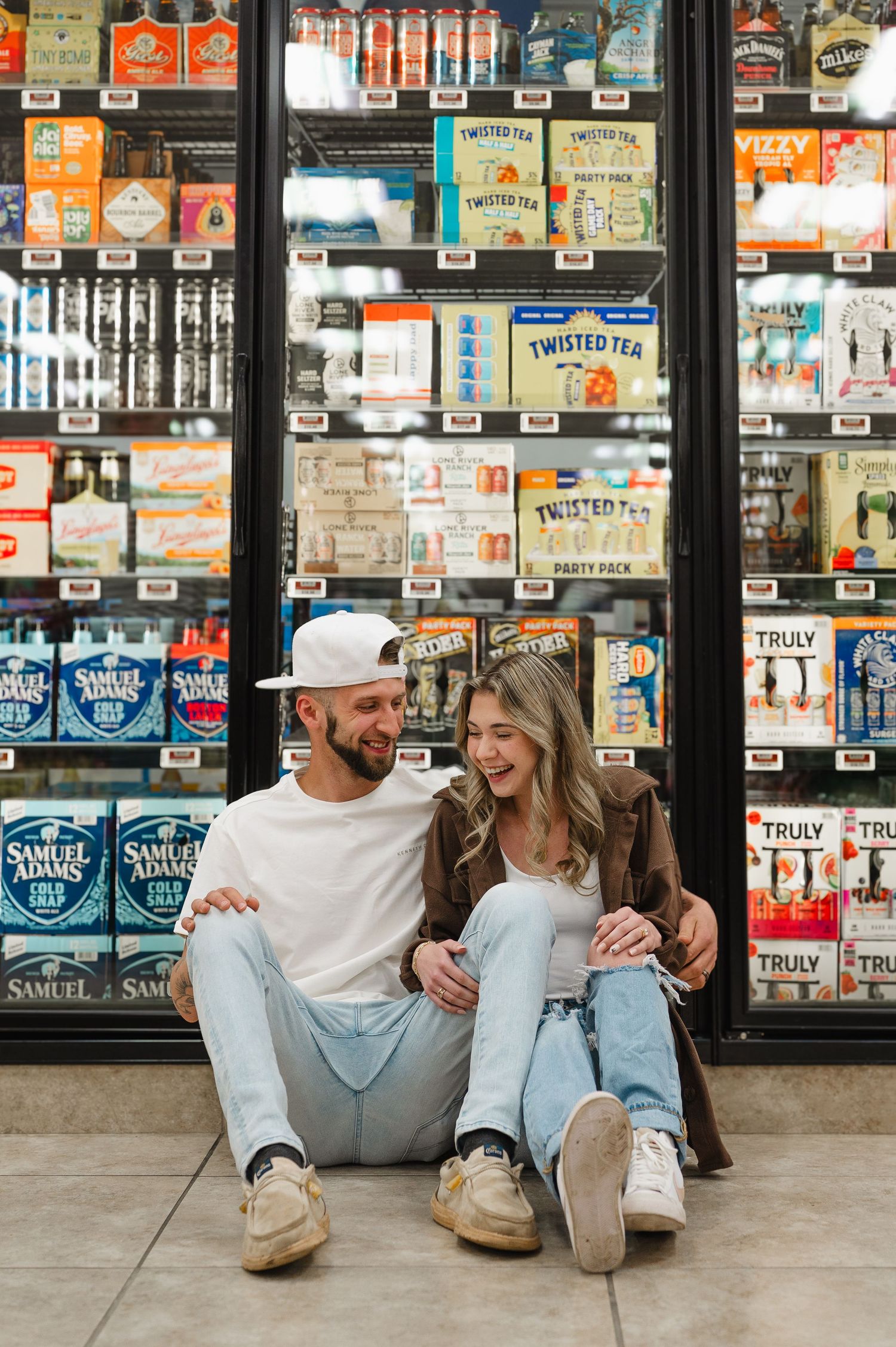 Couple sits on floor in front of convenience store refrigerator cases filled with beverages and drinks.