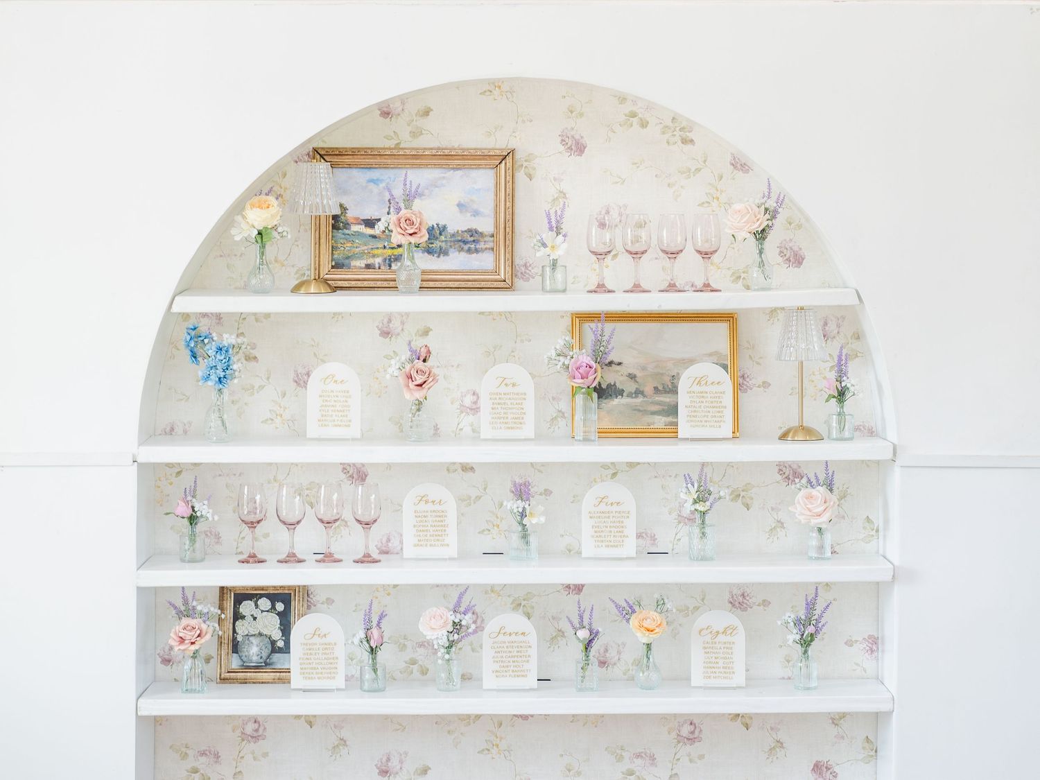A set of white floating shelves displaying wine glasses, bottles, and decorative items on a light-colored wall.