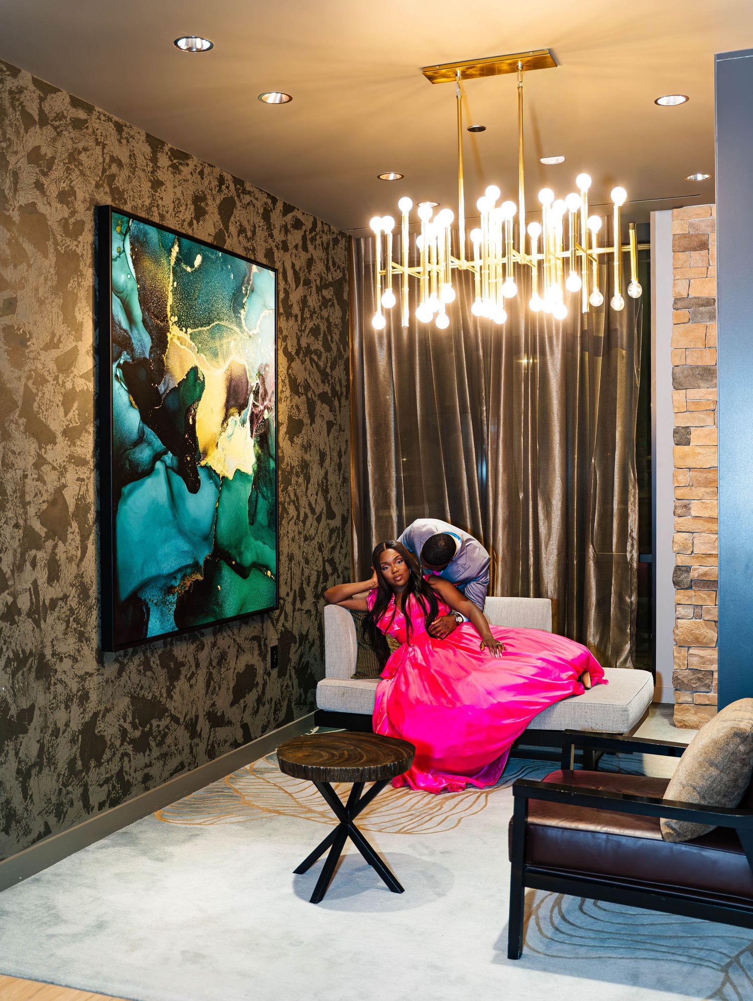 Woman in pink gown seated on white sofa in elegant room with abstract art, gold chandelier, and textured wallpaper.