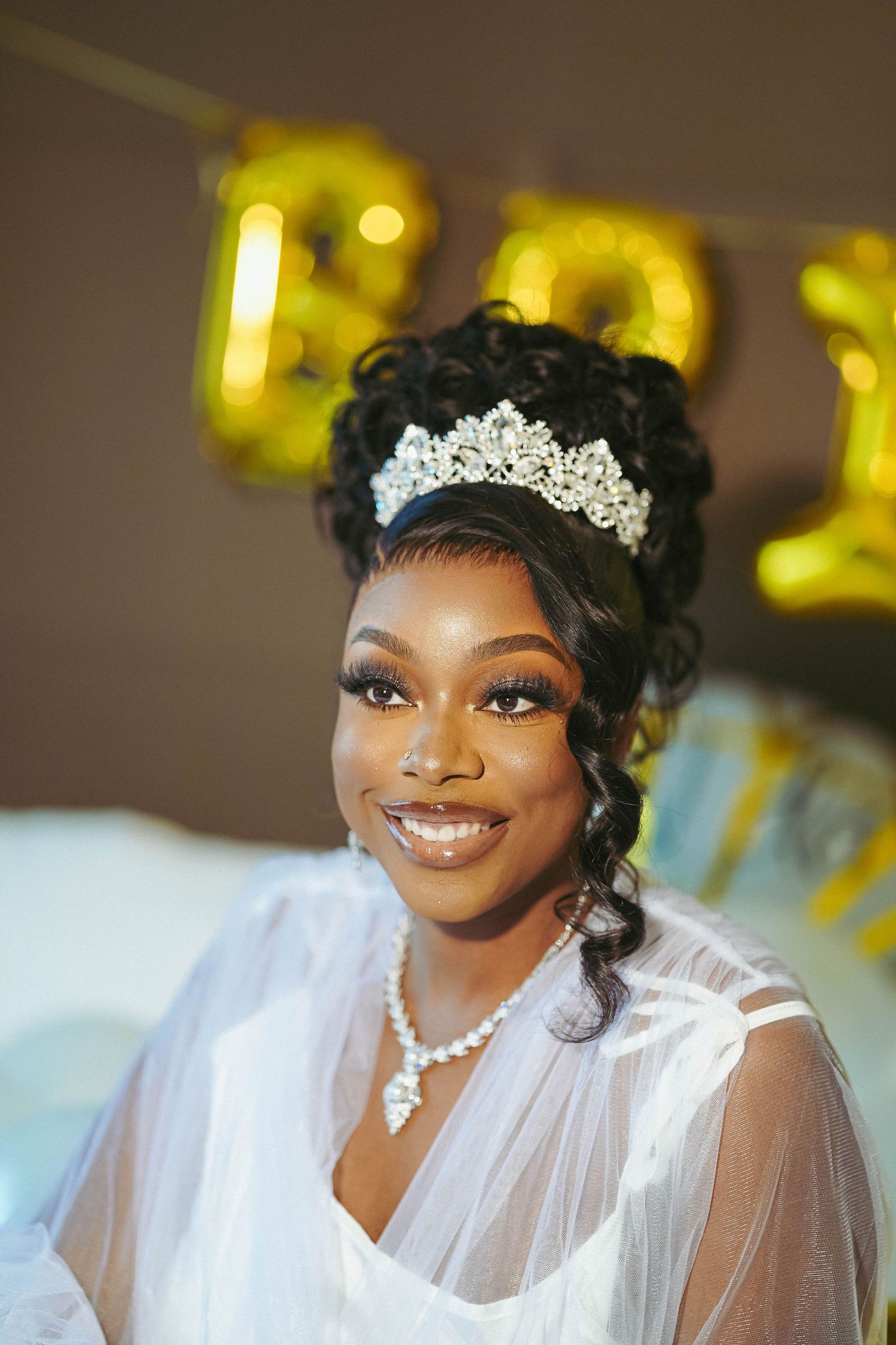 A bride wearing an elegant tiara and veil smiles radiantly in warm ambient lighting.